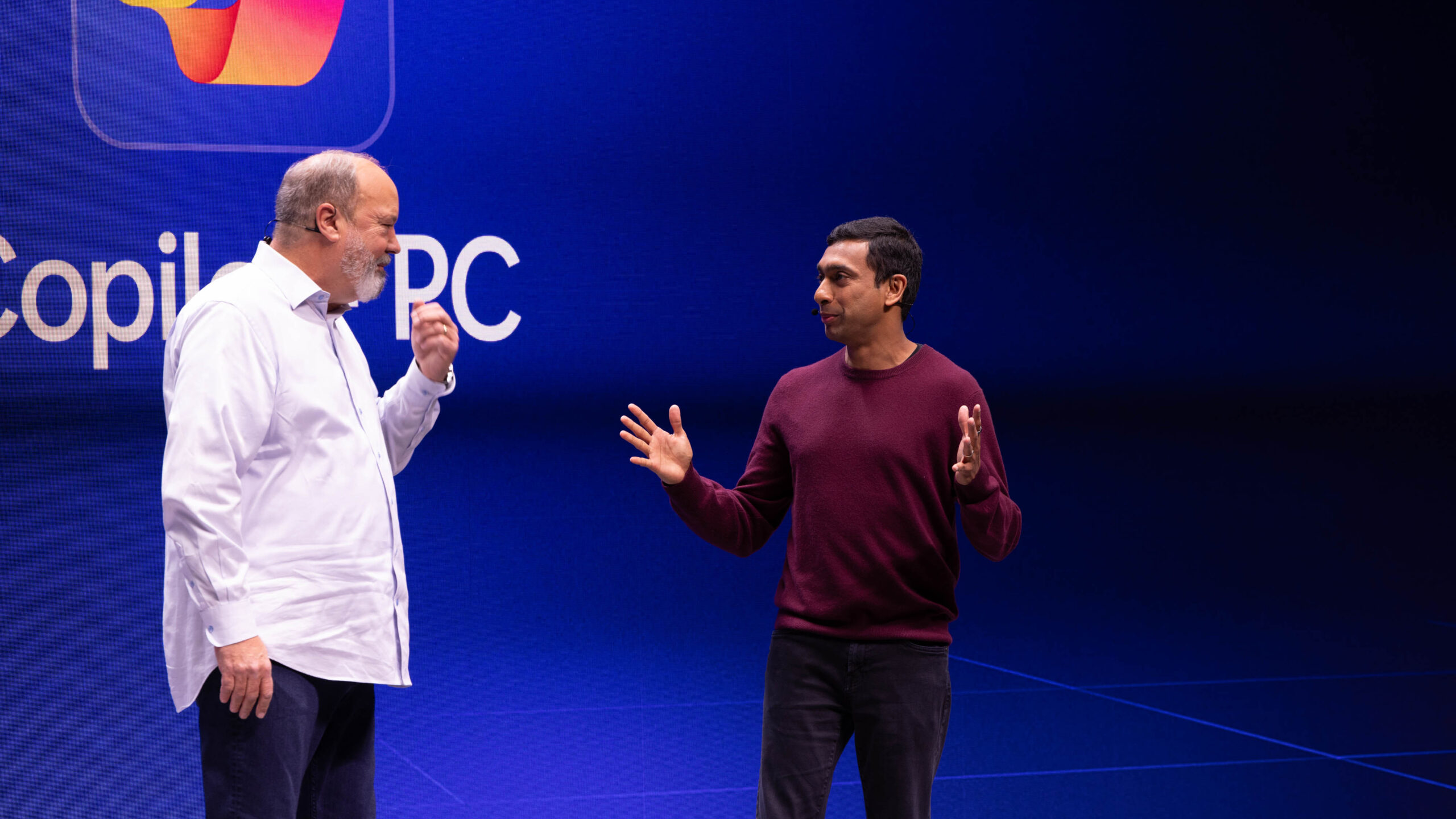 Two men are standing on a stage, having a discussion. The one on the left is wearing a white shirt, and the one on the right is wearing a maroon sweater. The background features a Copilot PC logo on a blue screen.