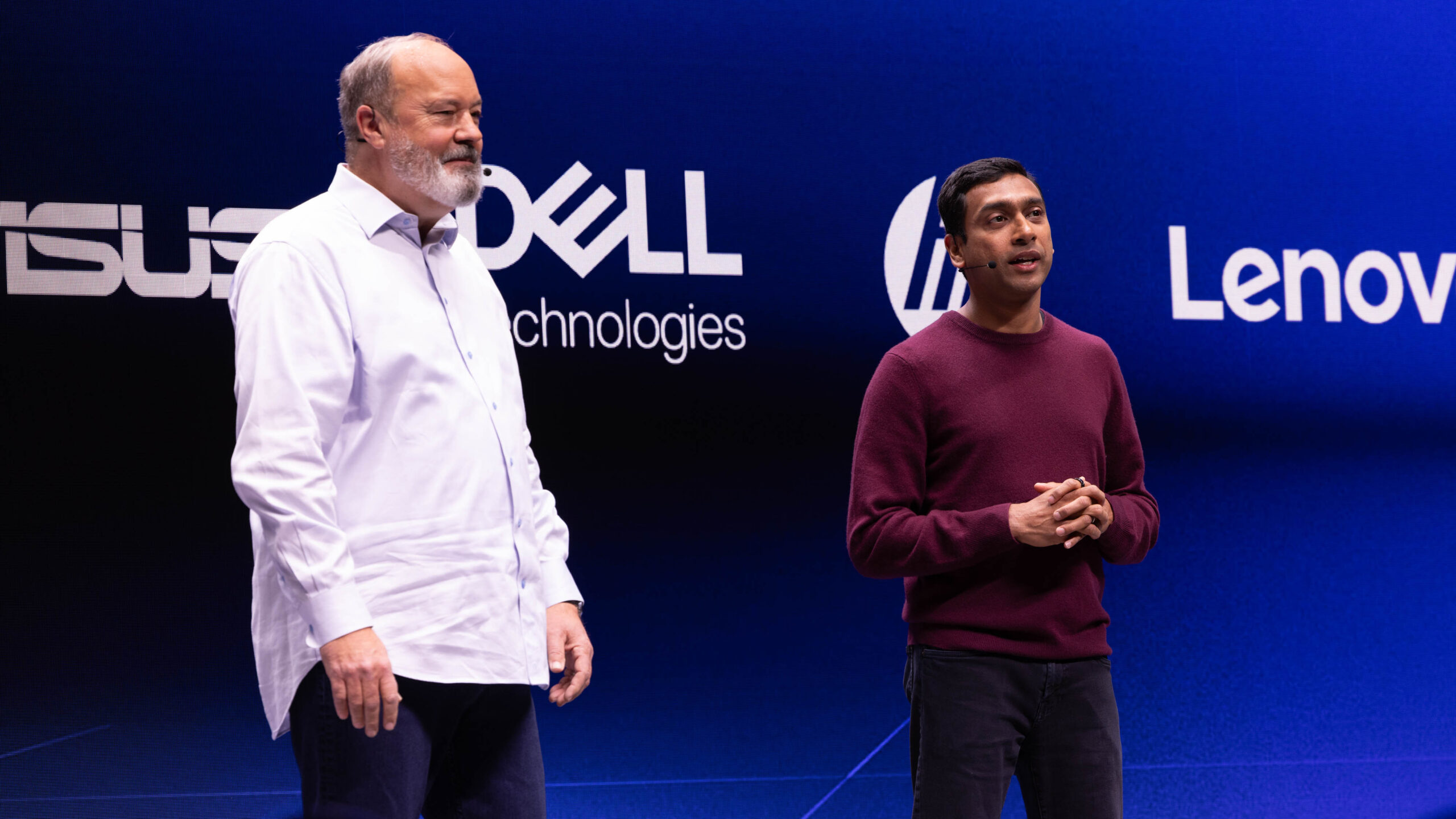 Two men stand on a stage with a blue background displaying logos of ASUS, Dell, Lenovo, and HP. One man wears a white shirt, and the other wears a maroon sweater. They appear to be speaking to an audience.