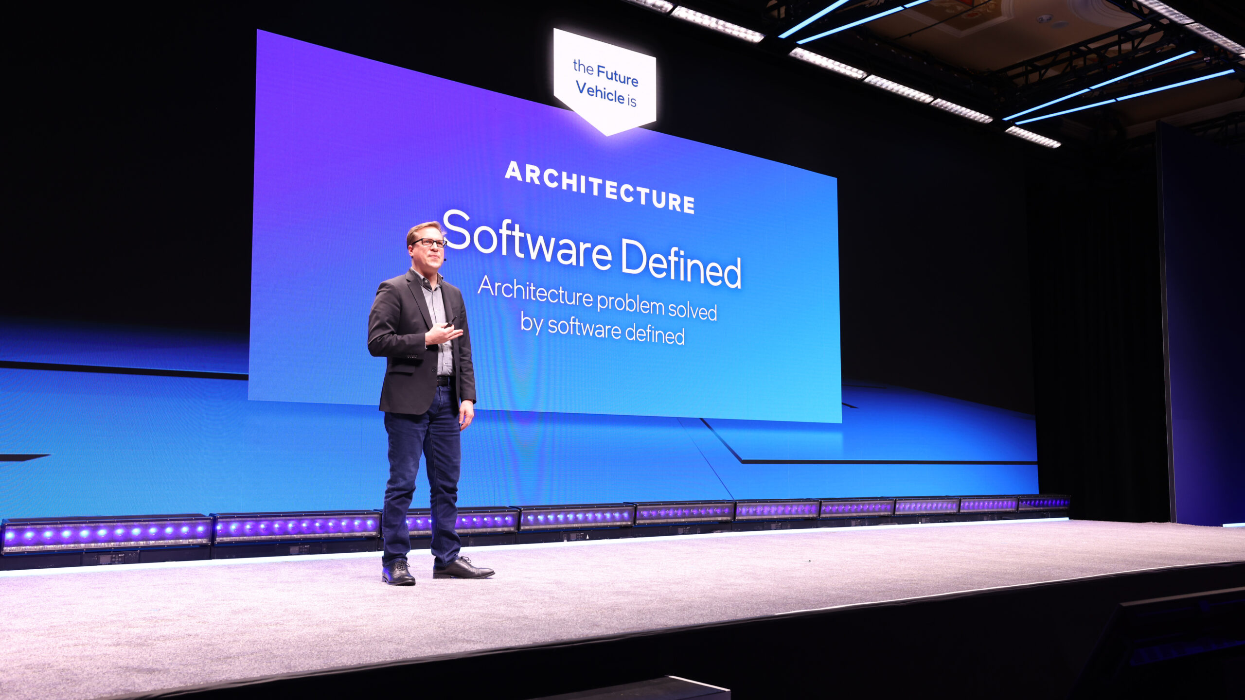 A person stands on a stage giving a presentation, with a large screen displaying the text ARCHITECTURE Software Defined. The stage is well-lit and the background features blue and purple colors.