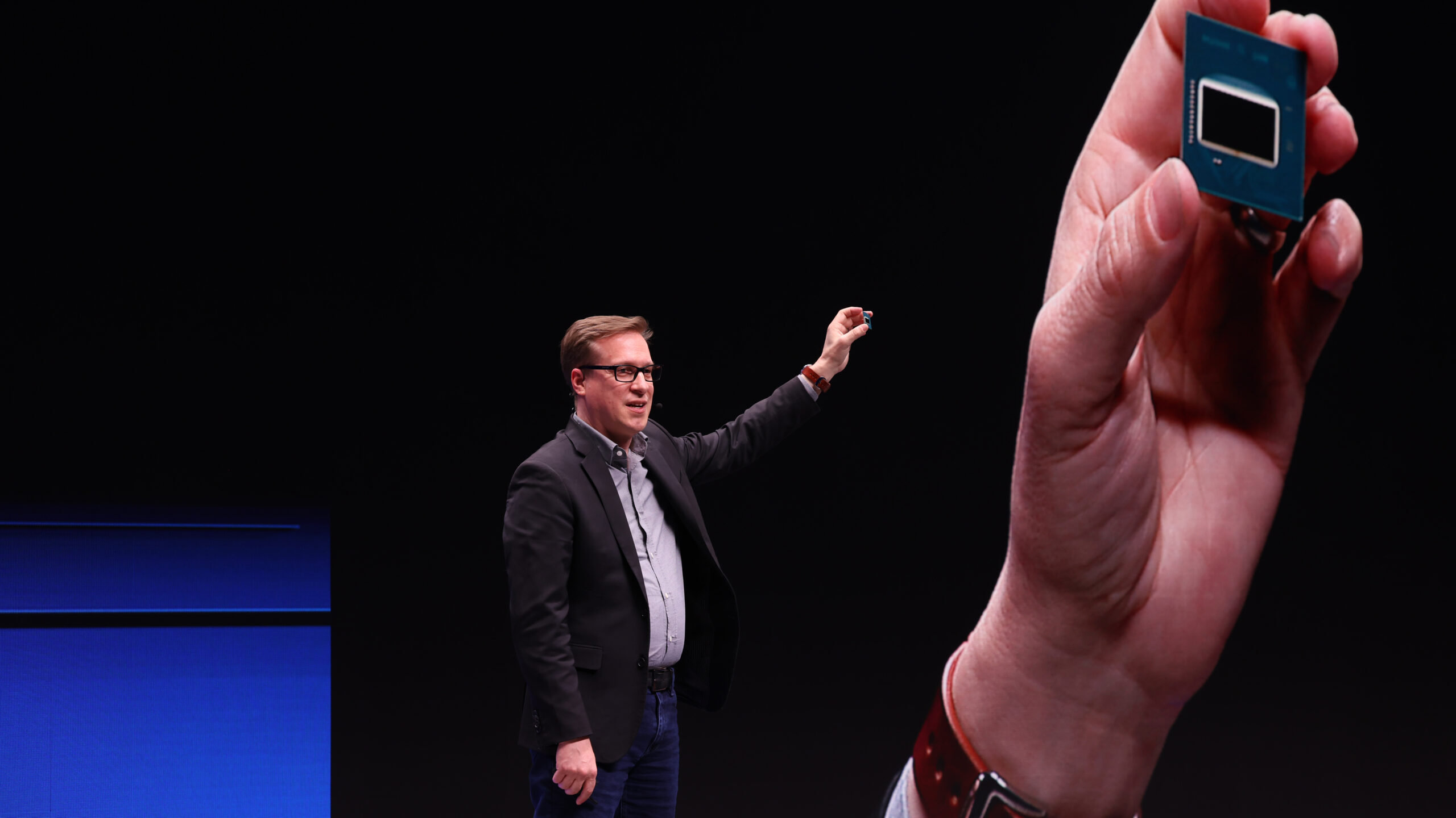A man in a dark suit and glasses speaks on stage, gesturing forward. In the foreground, a hand holds a small chip or processor against a dark background, highlighting its size.
