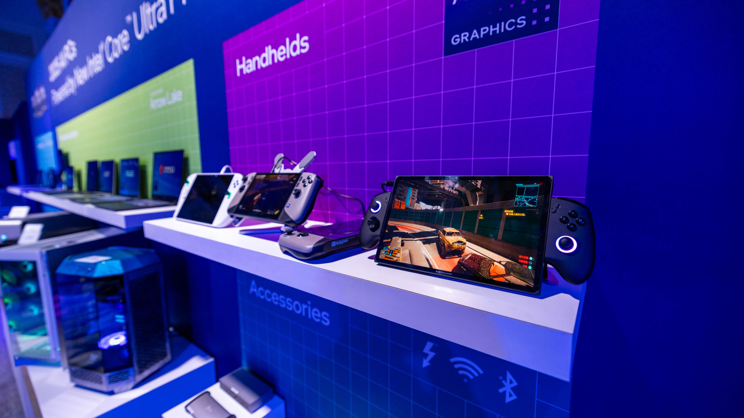 Two shelves display gaming devices and accessories at a tech expo. The upper shelf holds handheld gaming consoles, one running a racing game. The lower shelf shows various gaming accessories. The backdrop highlights product categories.