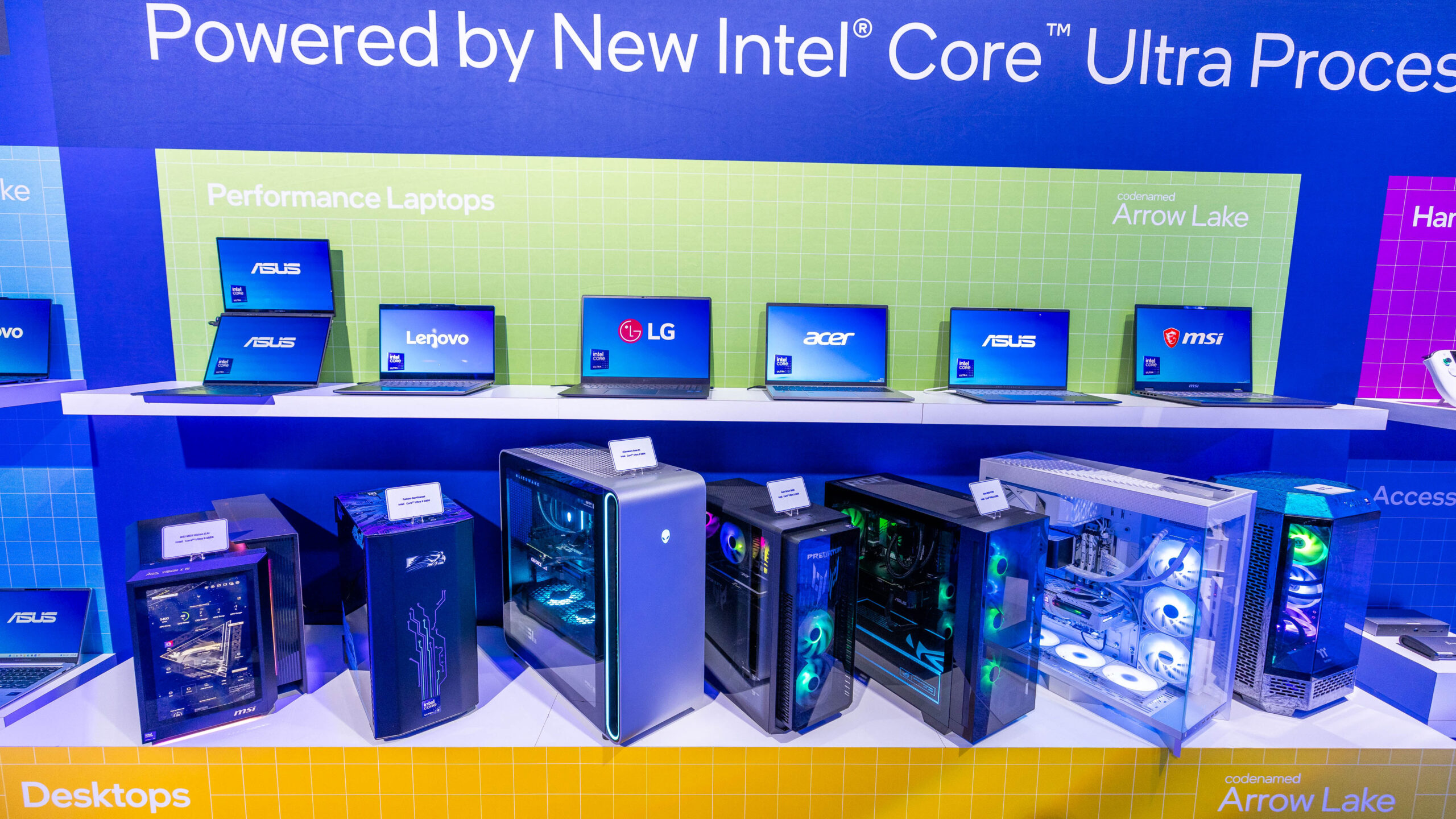 A display of desktop computers and laptops from brands like ASUS, Lenovo, LG, Acer, and MSI. The background reads Powered by New Intel Core Ultra Processors. The devices are showcased on white shelves with colorful lighting.