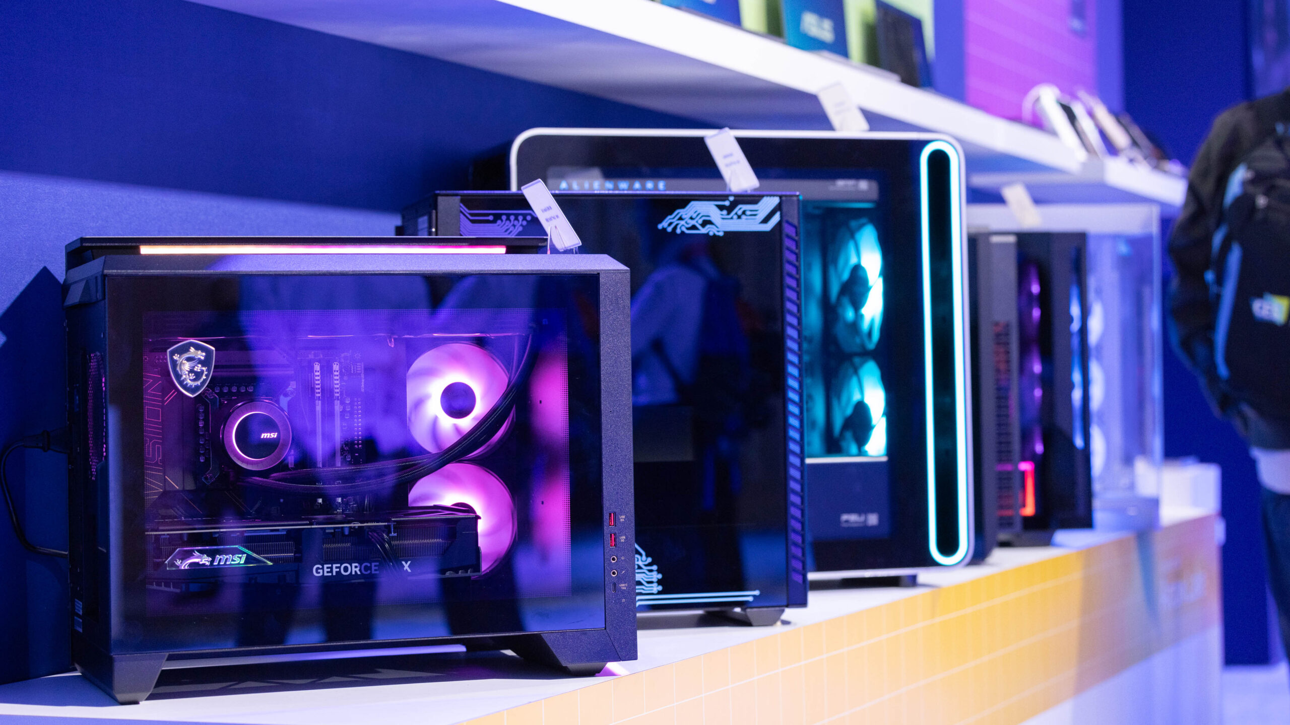 A row of modern gaming PCs displayed on a shelf. The first PC has a transparent side panel showing illuminated components with purple lighting. Additional PCs are lined up next to it, each featuring unique designs and lighting.