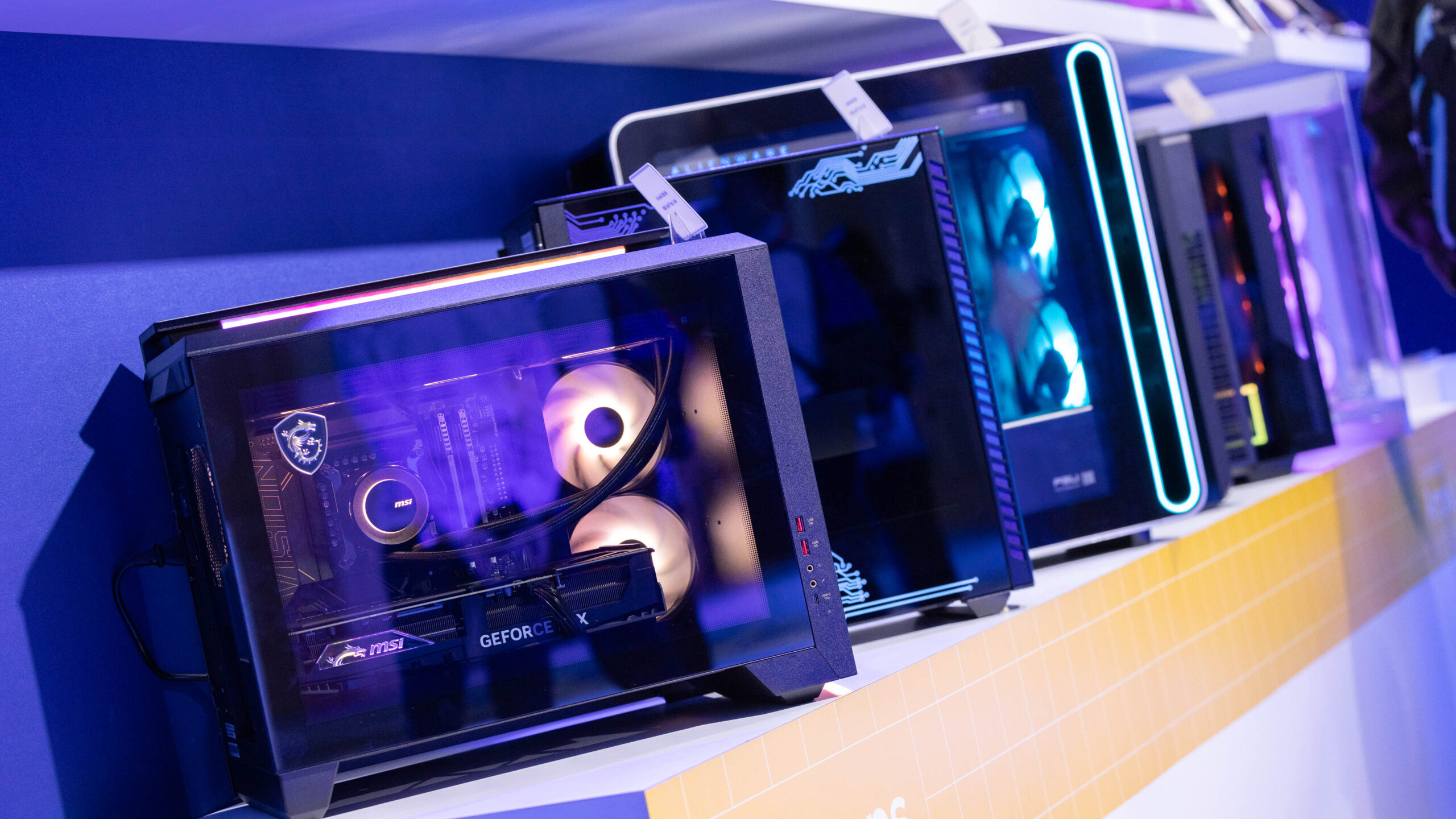 Row of gaming PCs on display, featuring transparent cases with internal RGB lighting, showcasing various components. Each computer is positioned on a shelf against a blue and yellow backdrop.