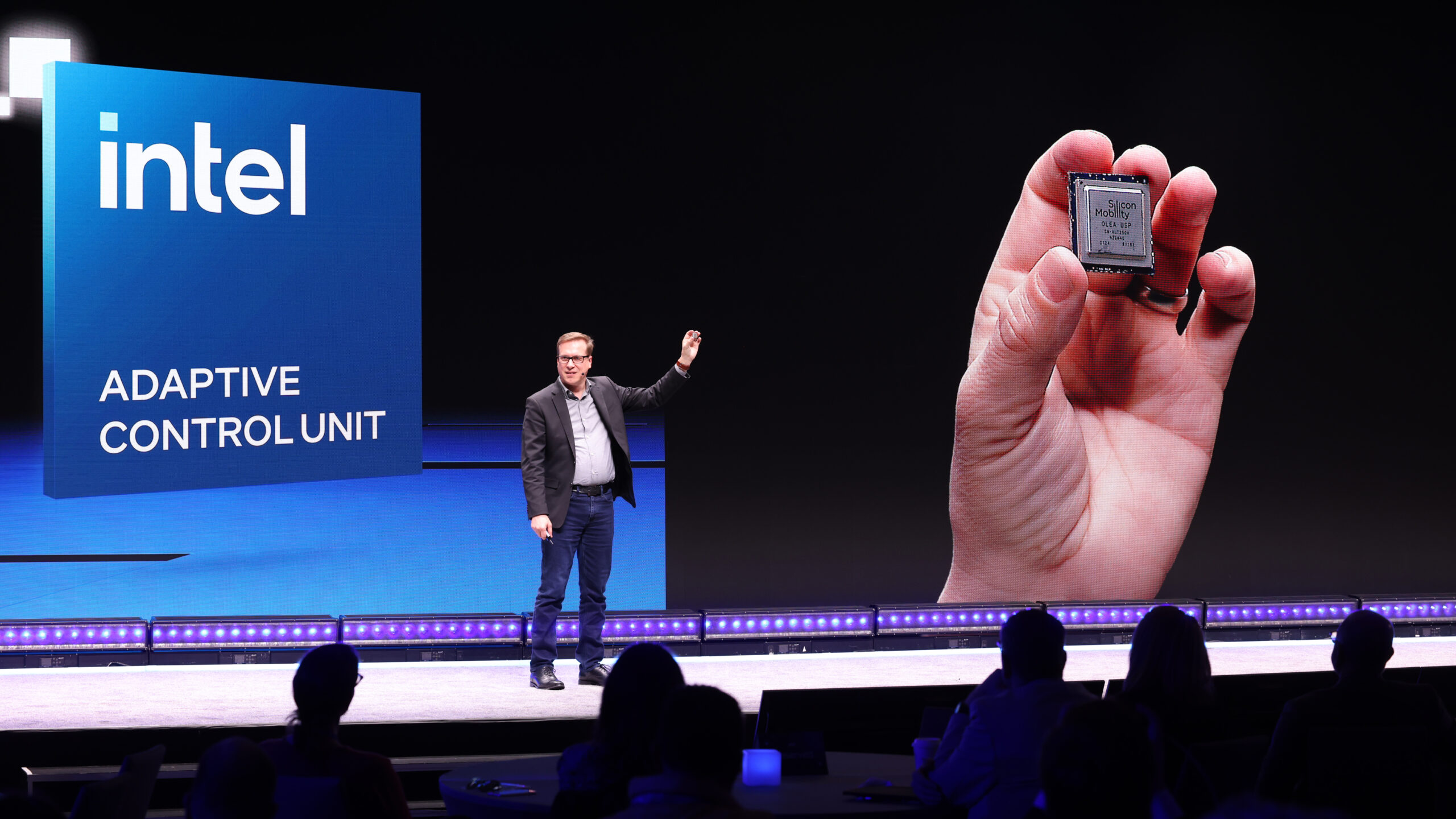 A speaker on stage presents the Intel Adaptive Control Unit, with a large screen showing a close-up of a hand holding the device. The background features the Intel logo and text reading ADAPTIVE CONTROL UNIT. Audience members are silhouetted in front.