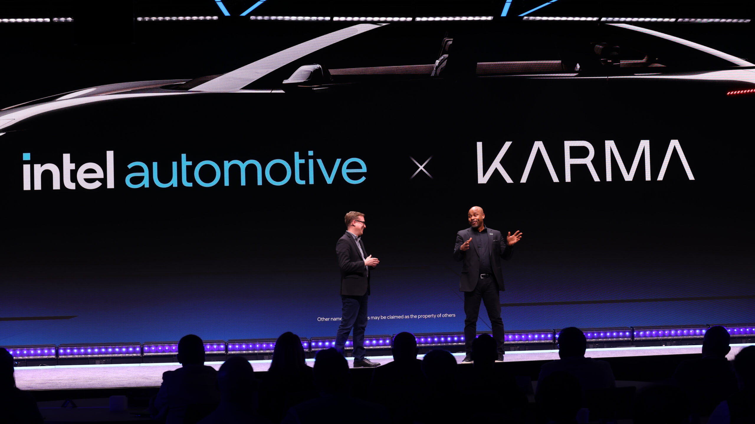 Two people are on stage presenting with a large screen behind them displaying intel automotive x KARMA, featuring the silhouettes of a car. The audience is seated in front of the stage in a dimly lit venue.