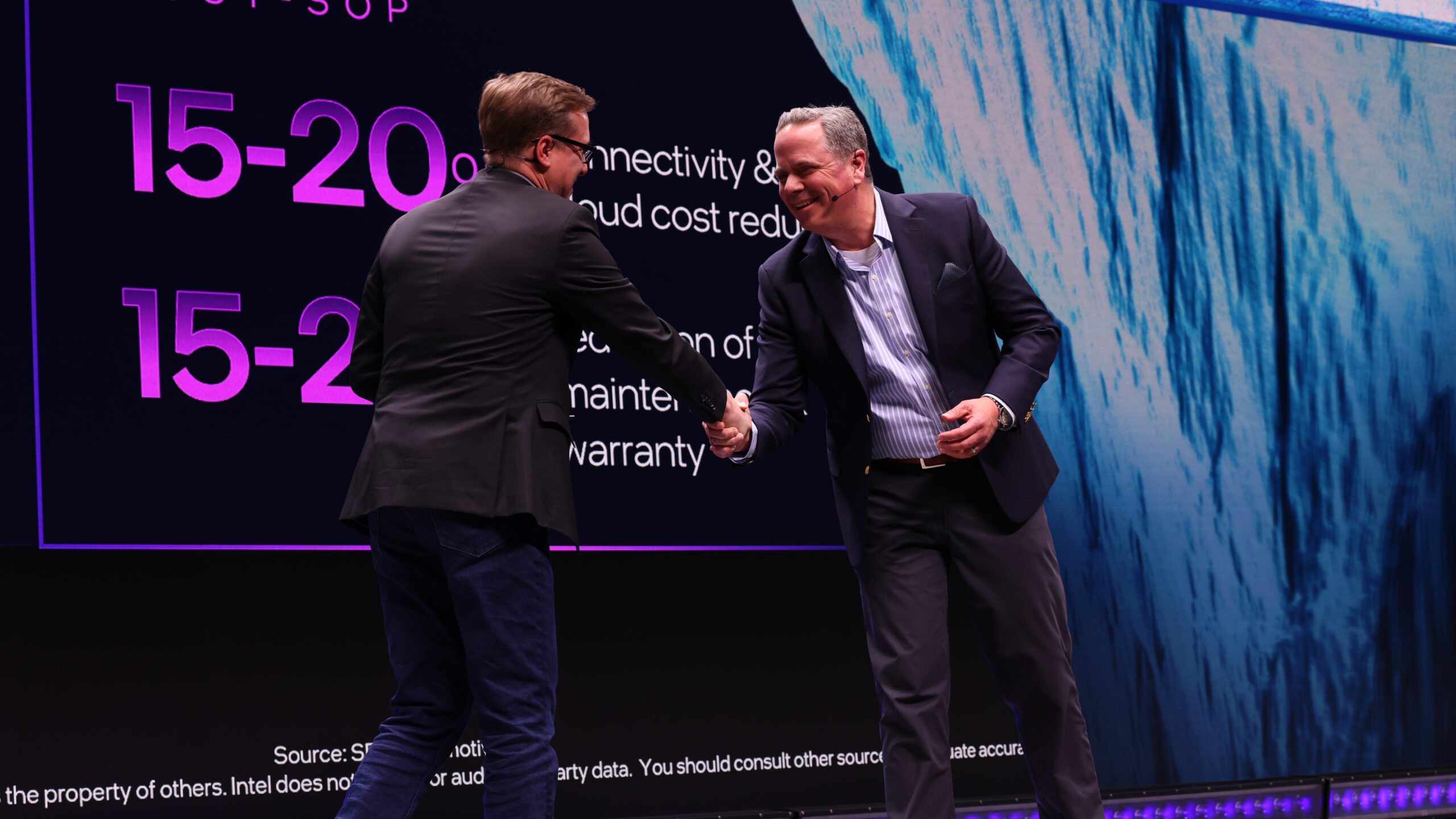 Two men in business attire shake hands on stage in front of a large screen displaying percentages. One man wears glasses and a dark suit, and the other is in a lighter suit. The background has a blend of blue patterns and text.