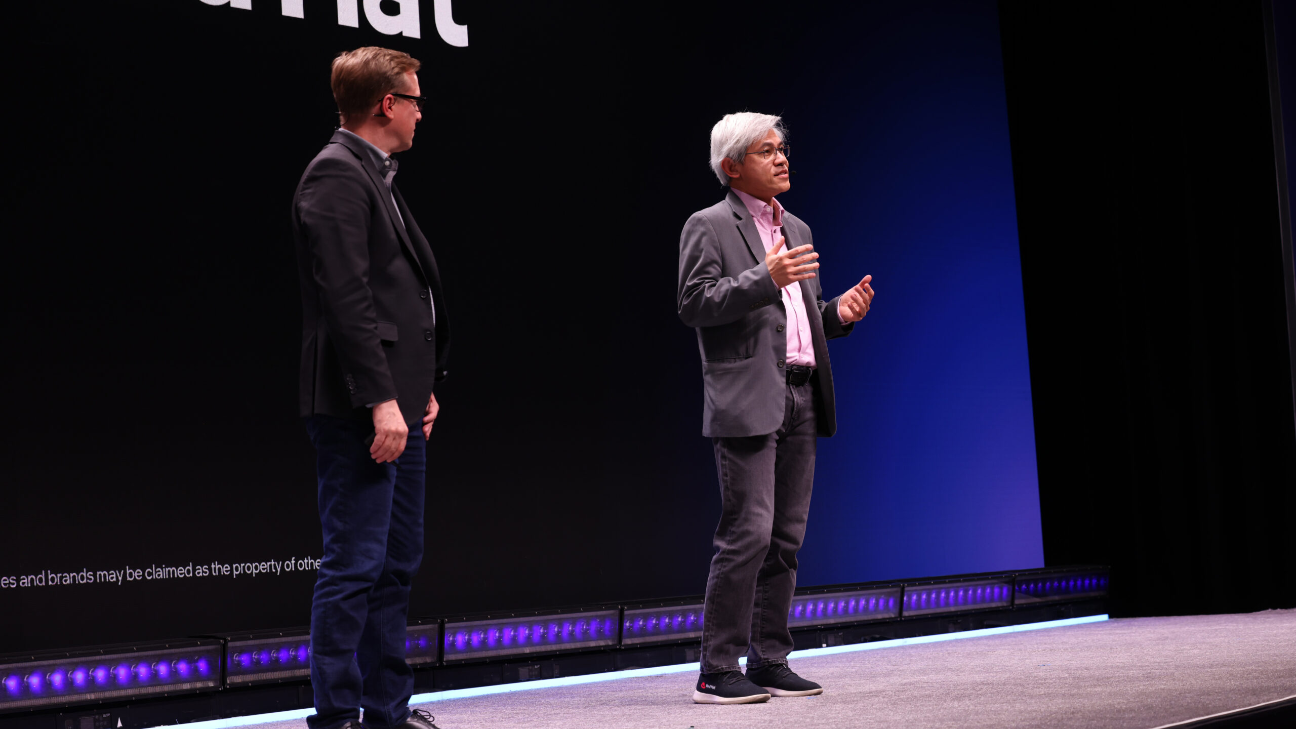 Two people stand on stage, engaged in conversation. One person is speaking while the other listens. Both are wearing jackets and jeans. The background is a large screen with partial text visible.