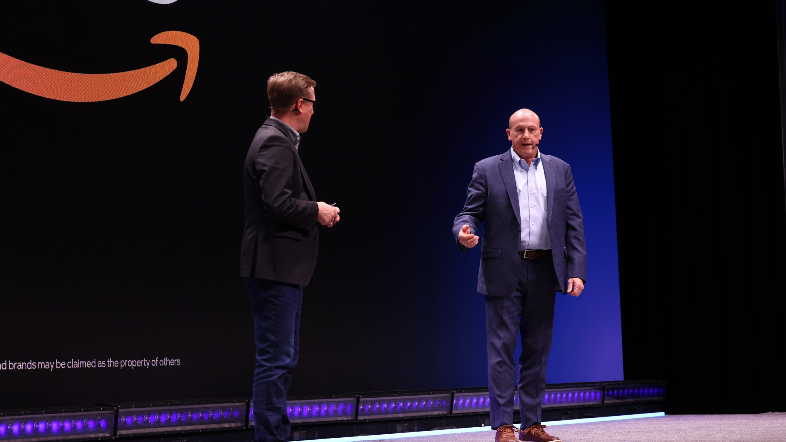 Two men are on stage in front of a large screen with an Amazon logo. One is in a black suit, the other in a blue suit. They appear to be having a discussion or presentation.