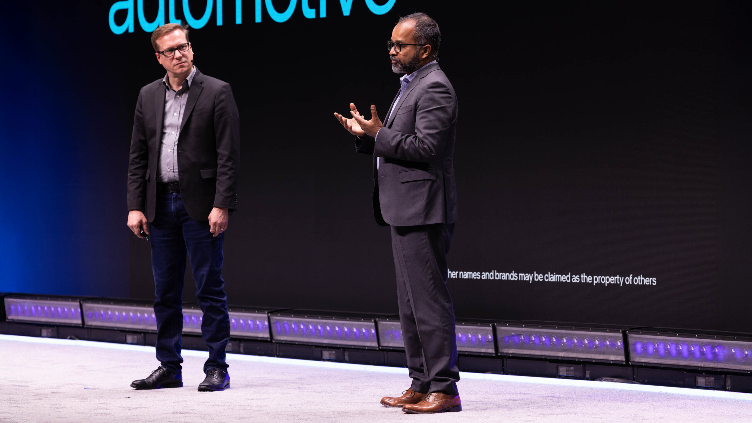 Two men in suits are standing on a stage with a large screen behind them displaying the word automotive. One man is speaking, while the other listens. The stage is lit with blue and purple lights.