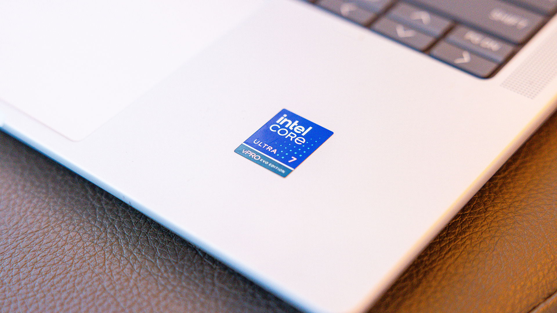 Close-up of a laptops corner showing an Intel Core Ultra i7 sticker on a metallic surface. The laptop is placed on a textured, brown surface. Part of the keyboard is visible in the top right corner.