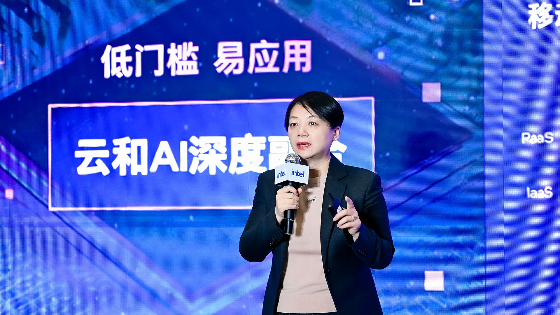 A person holding a microphone stands in front of a blue background with text in Chinese. They are wearing a dark suit and gesturing with their hand. The scene appears to be at a conference or presentation.