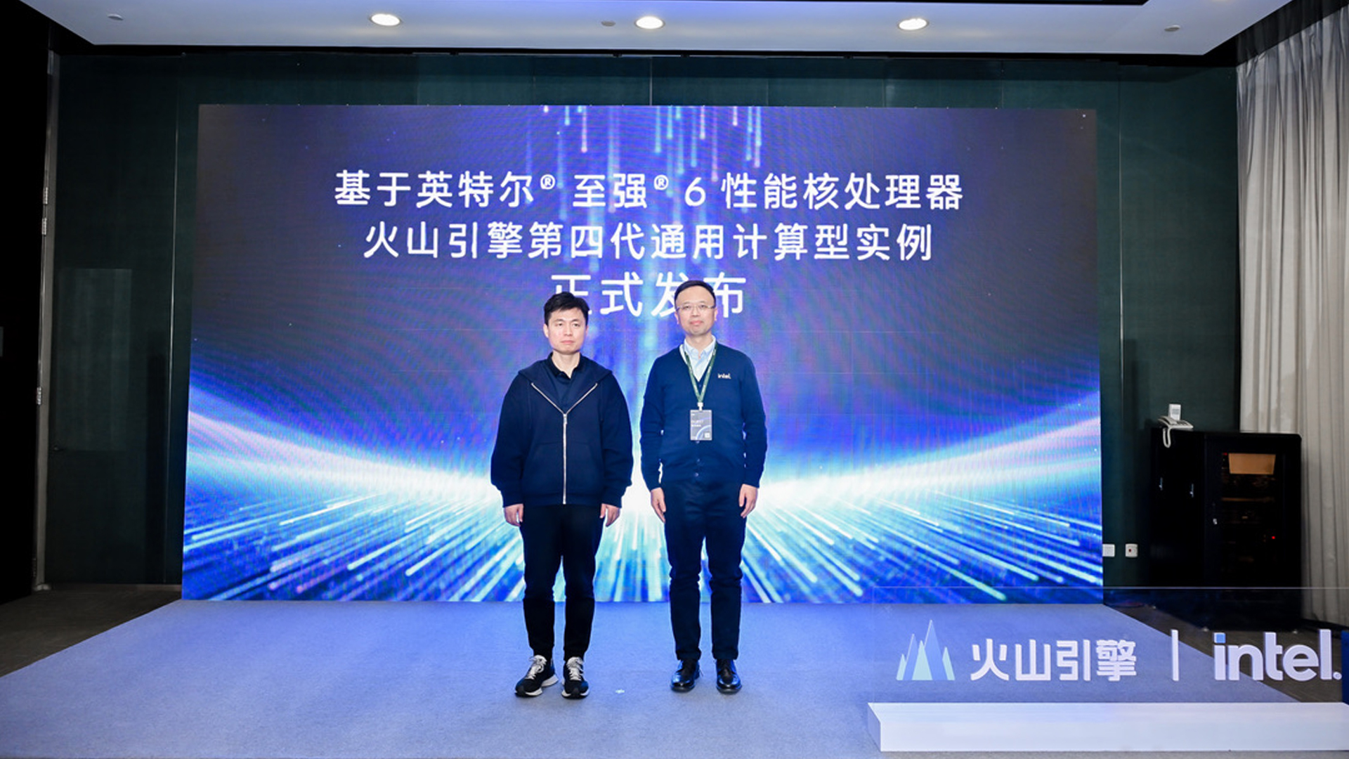 Two people stand on a stage in front of a large screen displaying text. The backdrop features graphics and logos of Intel and another brand. They are dressed in dark clothing, and the setting appears to be a formal presentation or event.