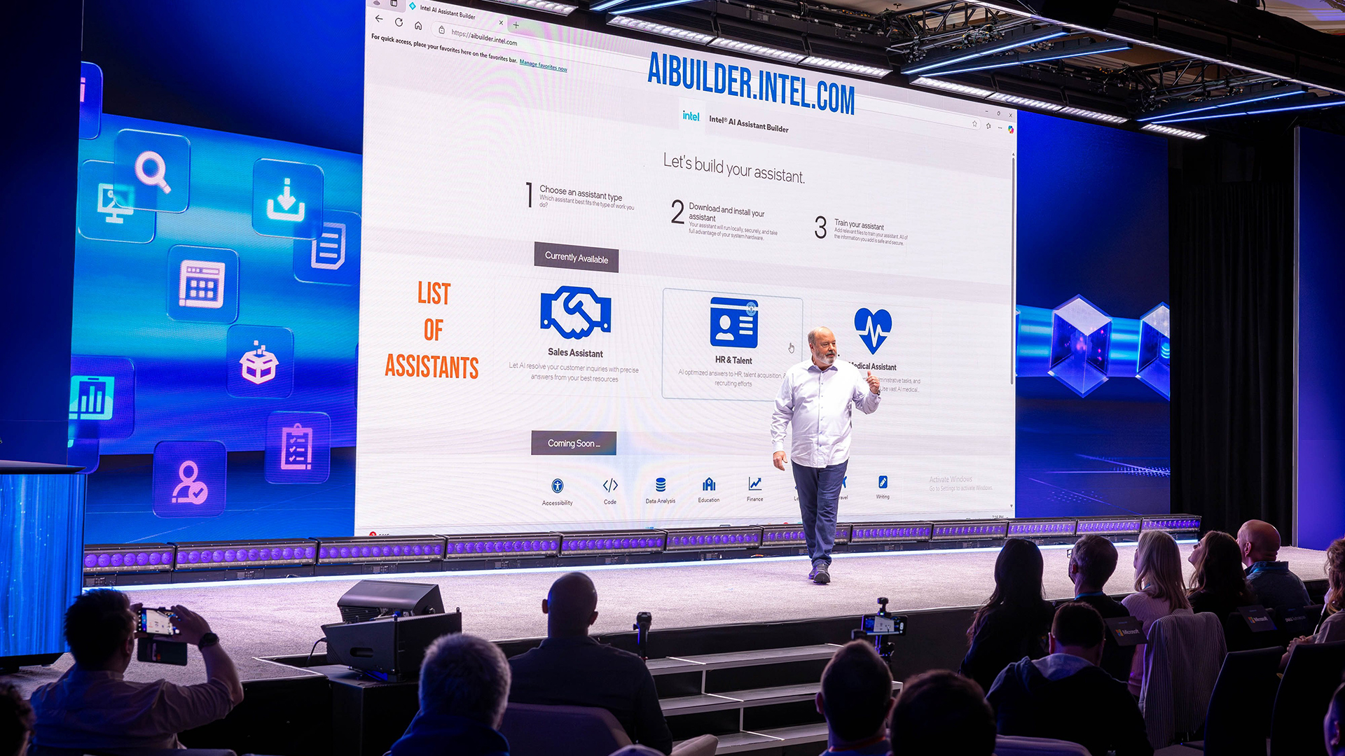 A man is presenting on a stage in front of a large screen displaying the website AIBUILDER.INTEL.COM with the title List of Assistants. The audience is seated in rows, with several cameras visible recording the event.
