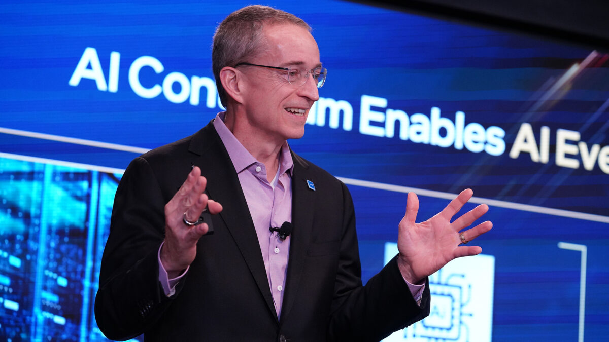 A person in a suit and glasses stands speaking in front of a large screen displaying text related to AI. They appear to be presenting or speaking at a conference or event.