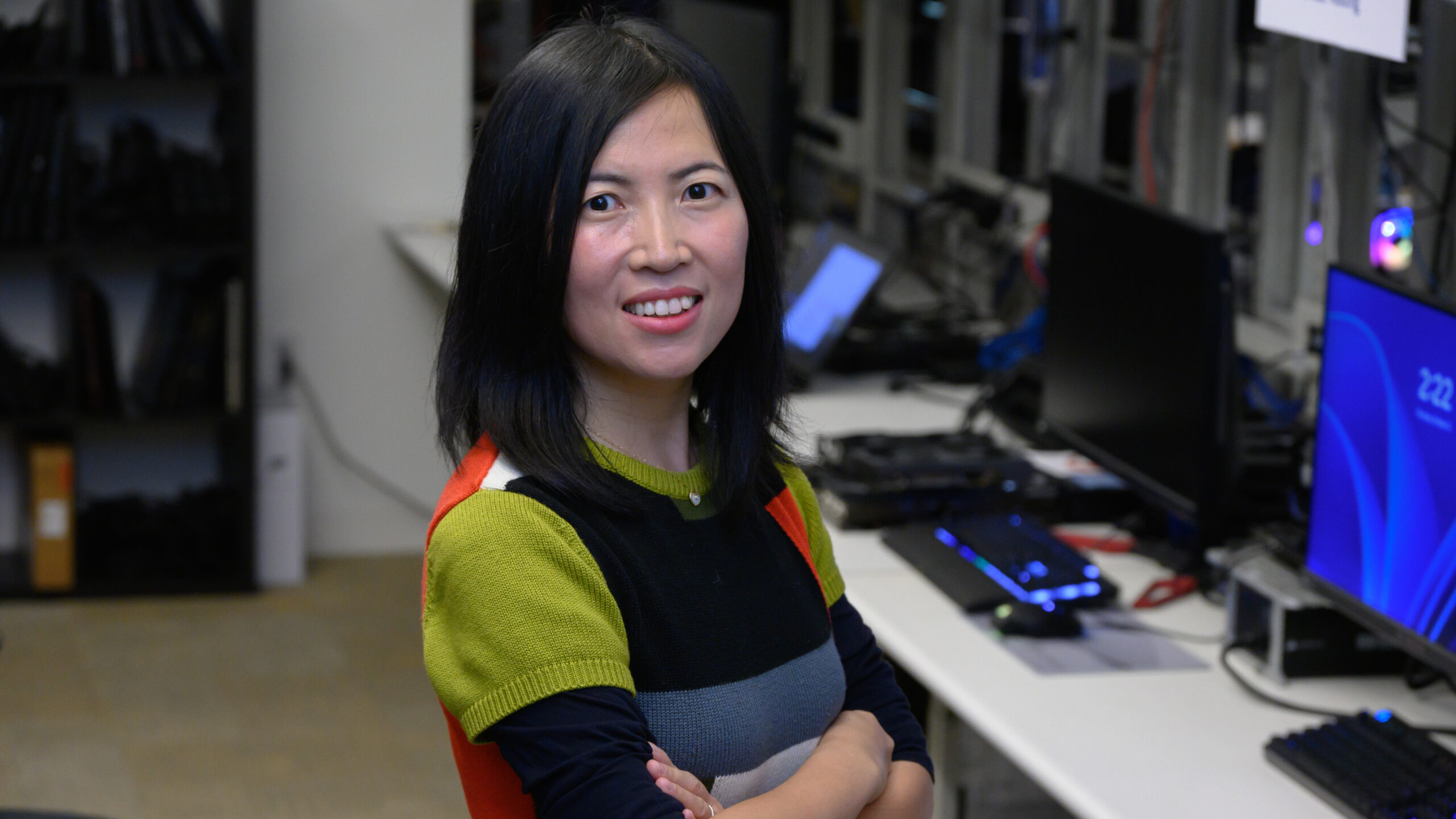A person with long black hair, wearing a colorful sweater, stands smiling with arms crossed in a room with computers and equipment in the background.