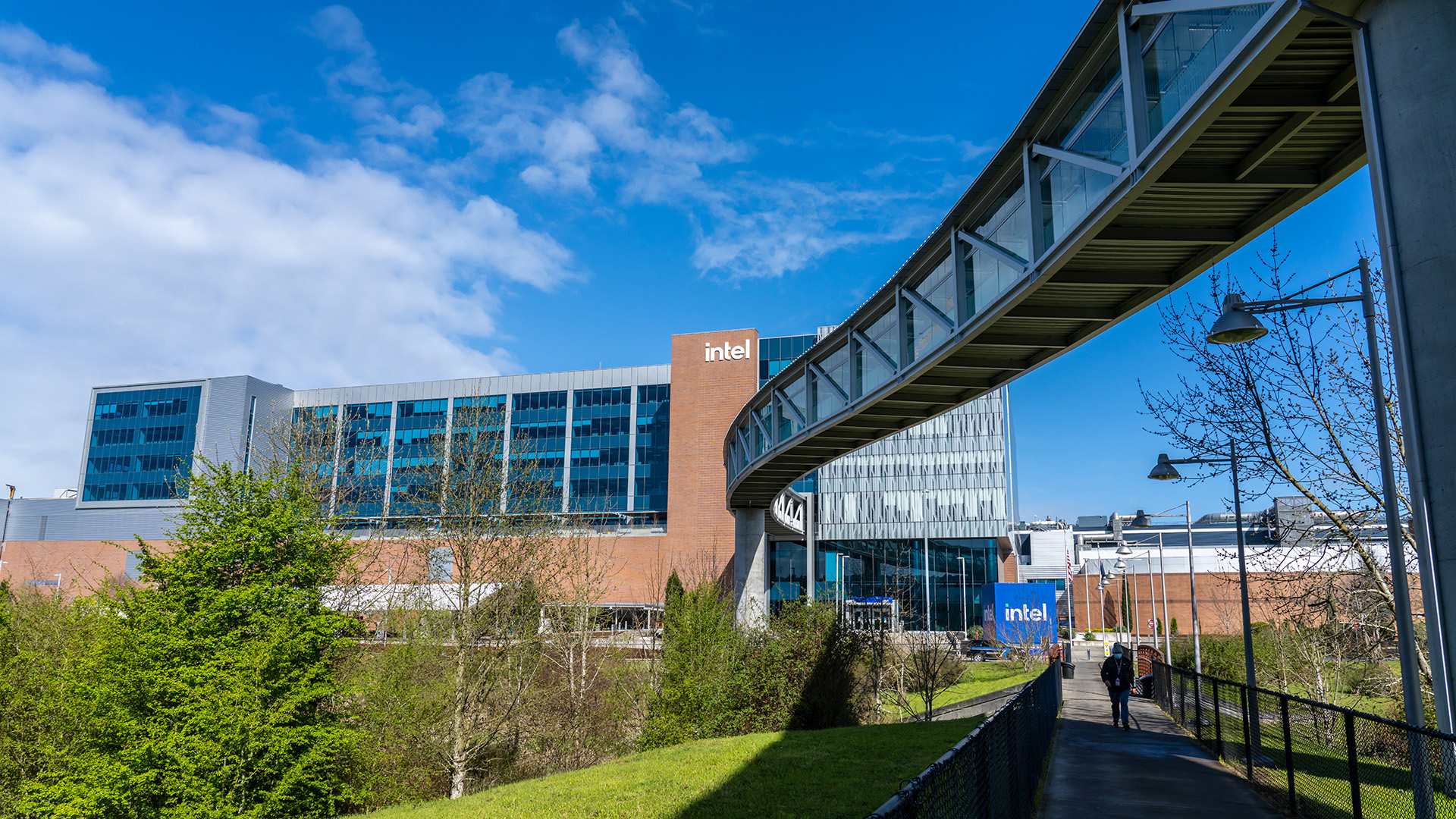 Exterior view of an Intel building with a blue sky above. A modern skybridge is visible, curving towards the building. Trees and a grassy area are in the foreground, with a pathway and people walking on it.
