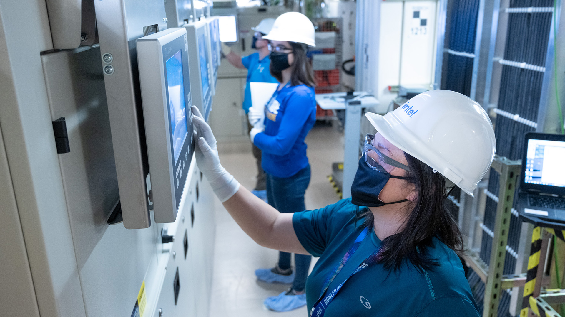 Three people in protective gear work in a laboratory. The foreground individual interacts with a touchscreen while others check equipment. Theyre all wearing helmets, gloves, and masks, emphasizing safety.