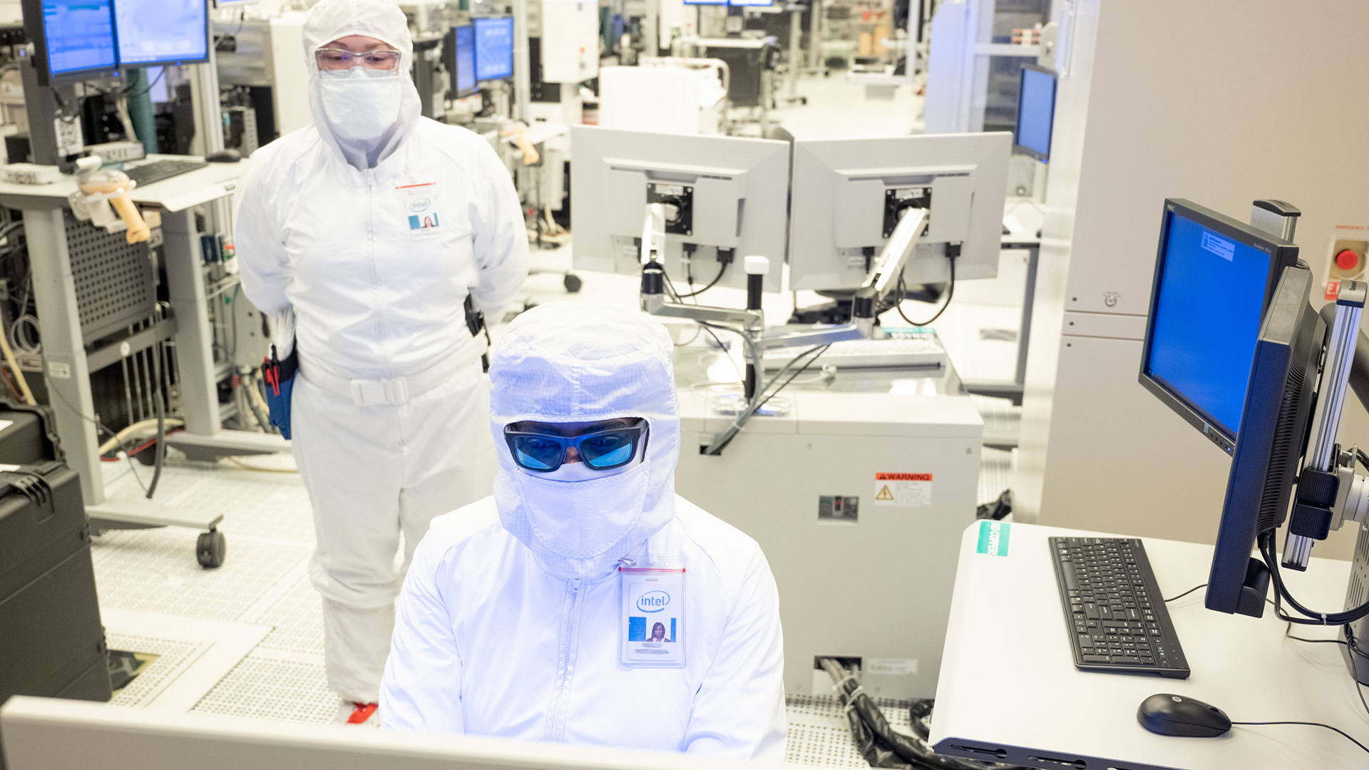 Two individuals in cleanroom suits and tinted goggles work in a laboratory. One is seated at a computer, while the other stands nearby. The room is filled with electronic equipment and monitors.