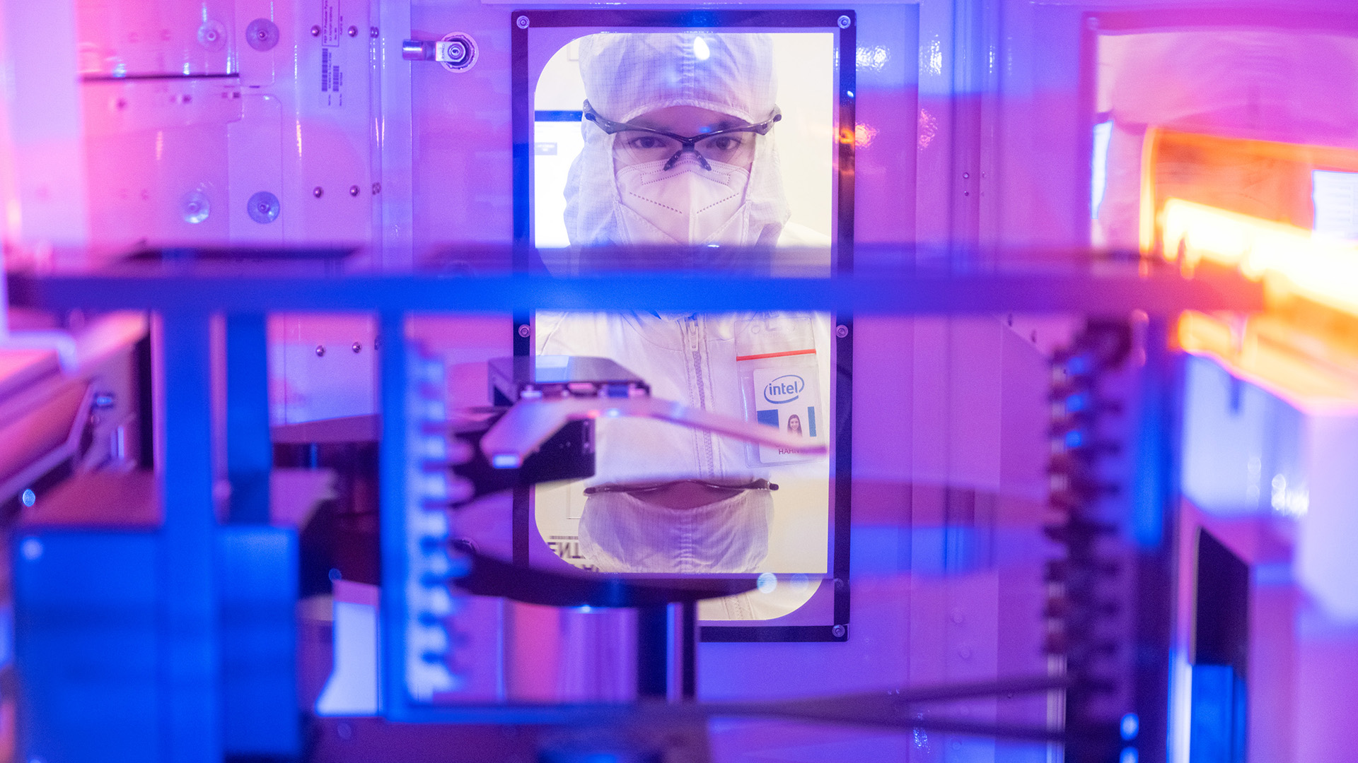 Person in protective lab suit and goggles seen through a window inside a tech lab, surrounded by machinery with blue and purple lighting.