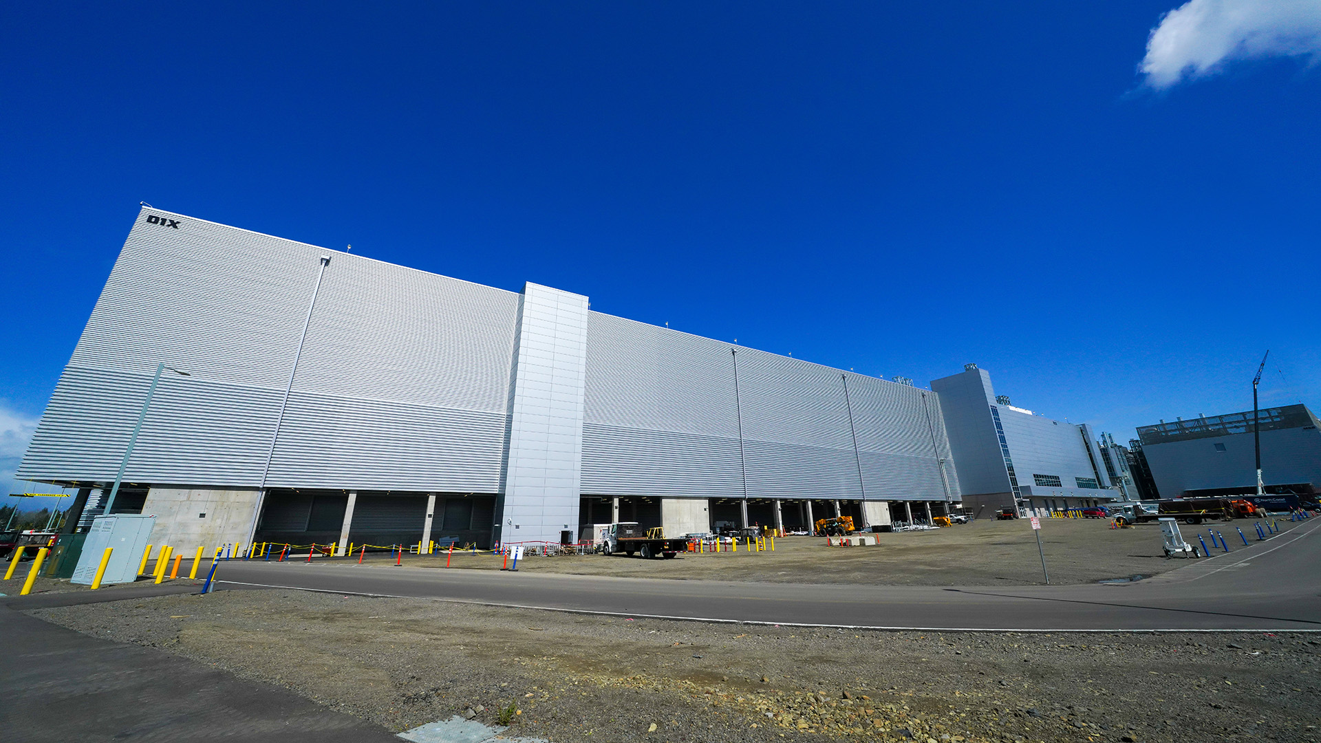 A large industrial building with a modern design stands under a clear blue sky. The pale gray structure has multiple levels and a textured facade. Construction equipment and vehicles are visible around the premises on a paved area.