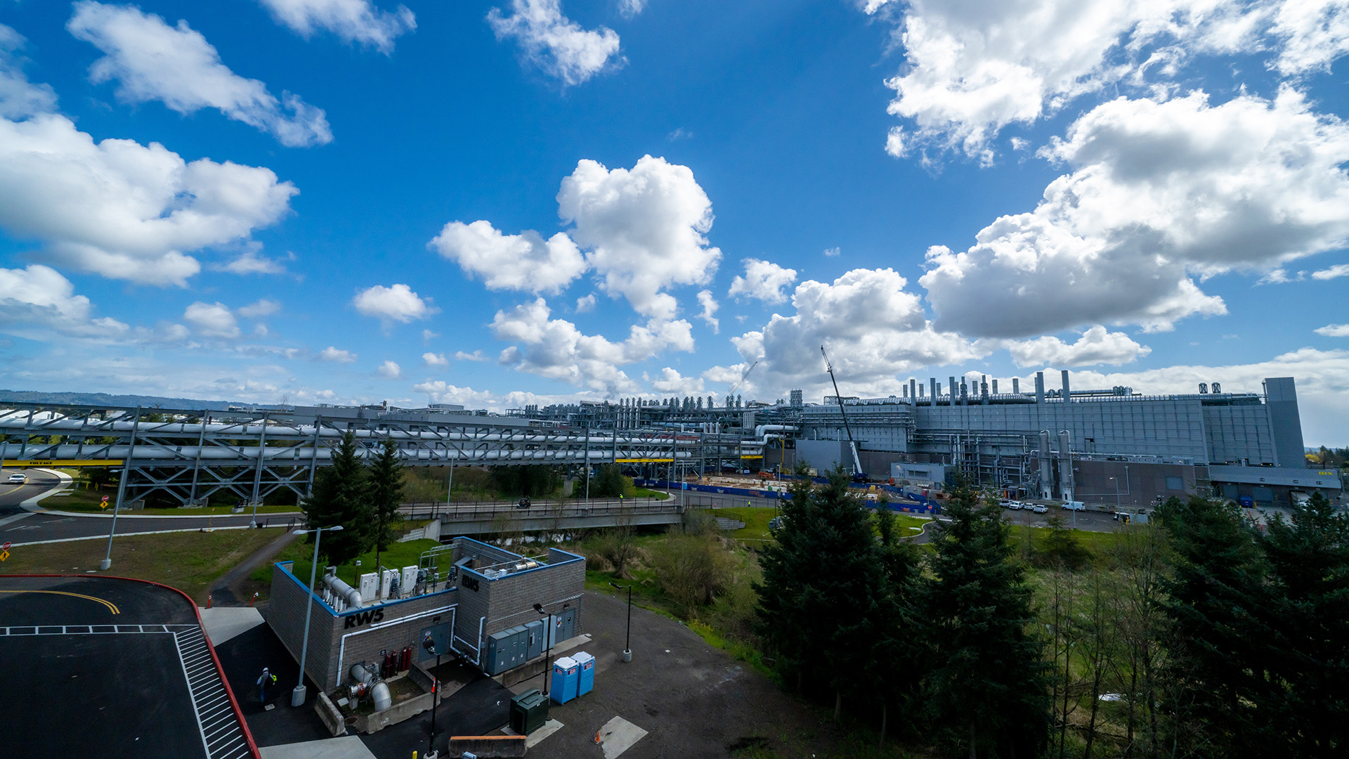 A large industrial complex with multiple buildings and chimneys under a blue sky with fluffy clouds. Trees and greenery are visible in the foreground. The facility has a street with a bridge and some utility structures nearby.