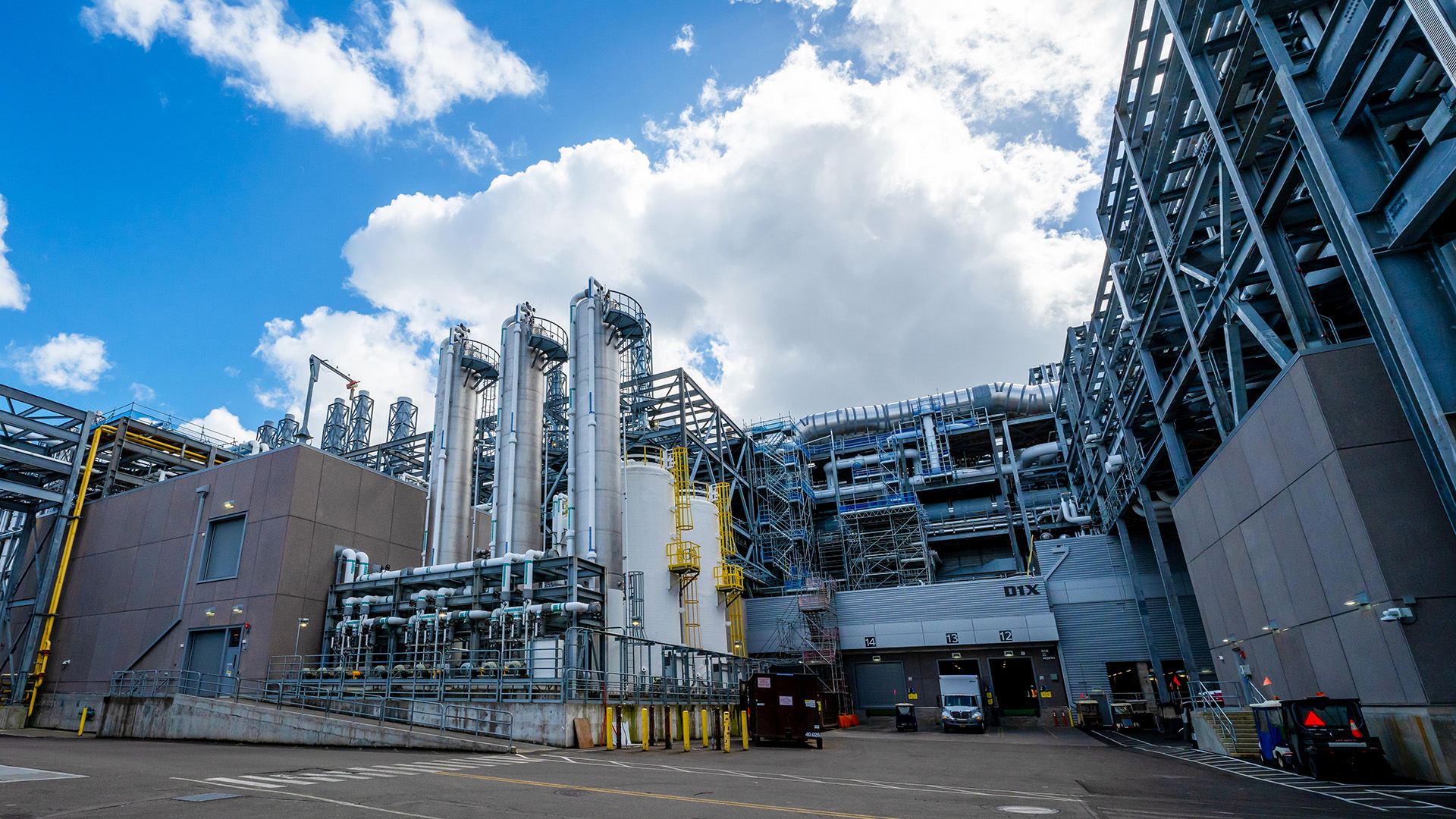 Industrial facility with large metal structures and pipes under a partly cloudy sky. Several tall cylindrical storage tanks are visible. Building marked with DIX on the side. Parking area in the foreground with a few vehicles.