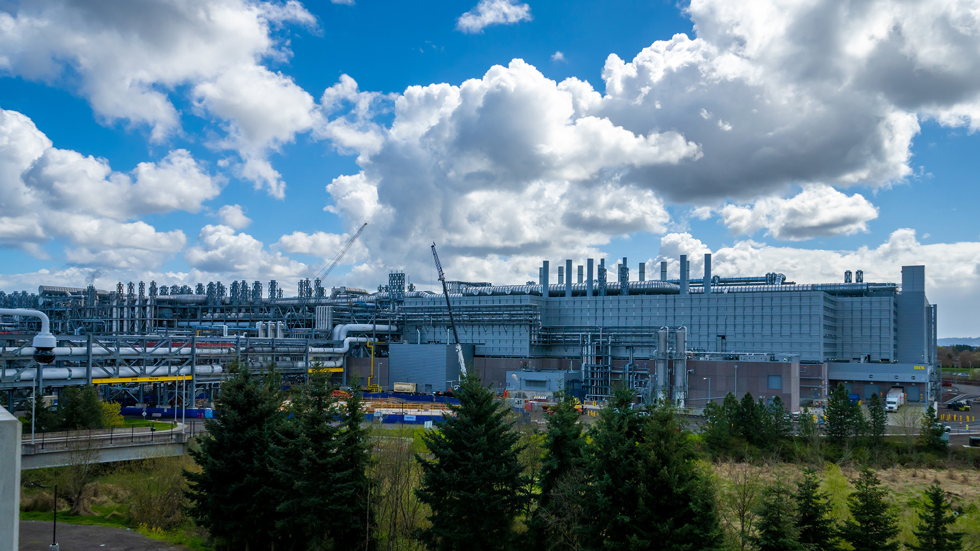 A large industrial facility with numerous chimneys and pipes is set against a partly cloudy sky. The foreground features trees and a grassy area. Cranes are visible, suggesting ongoing construction or maintenance.