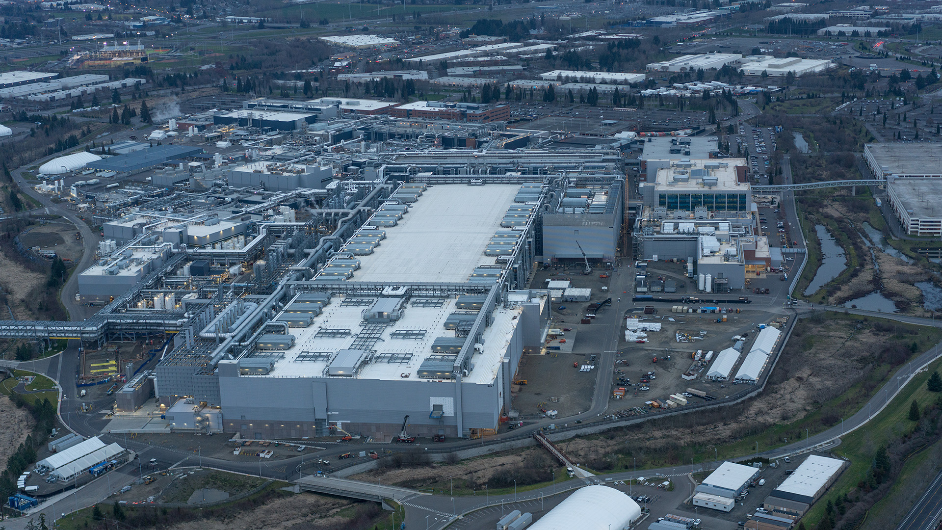 Aerial view of an industrial complex with large white-roofed buildings, surrounded by roads, parking areas, and smaller structures. The landscape includes greenery and patches of open land.