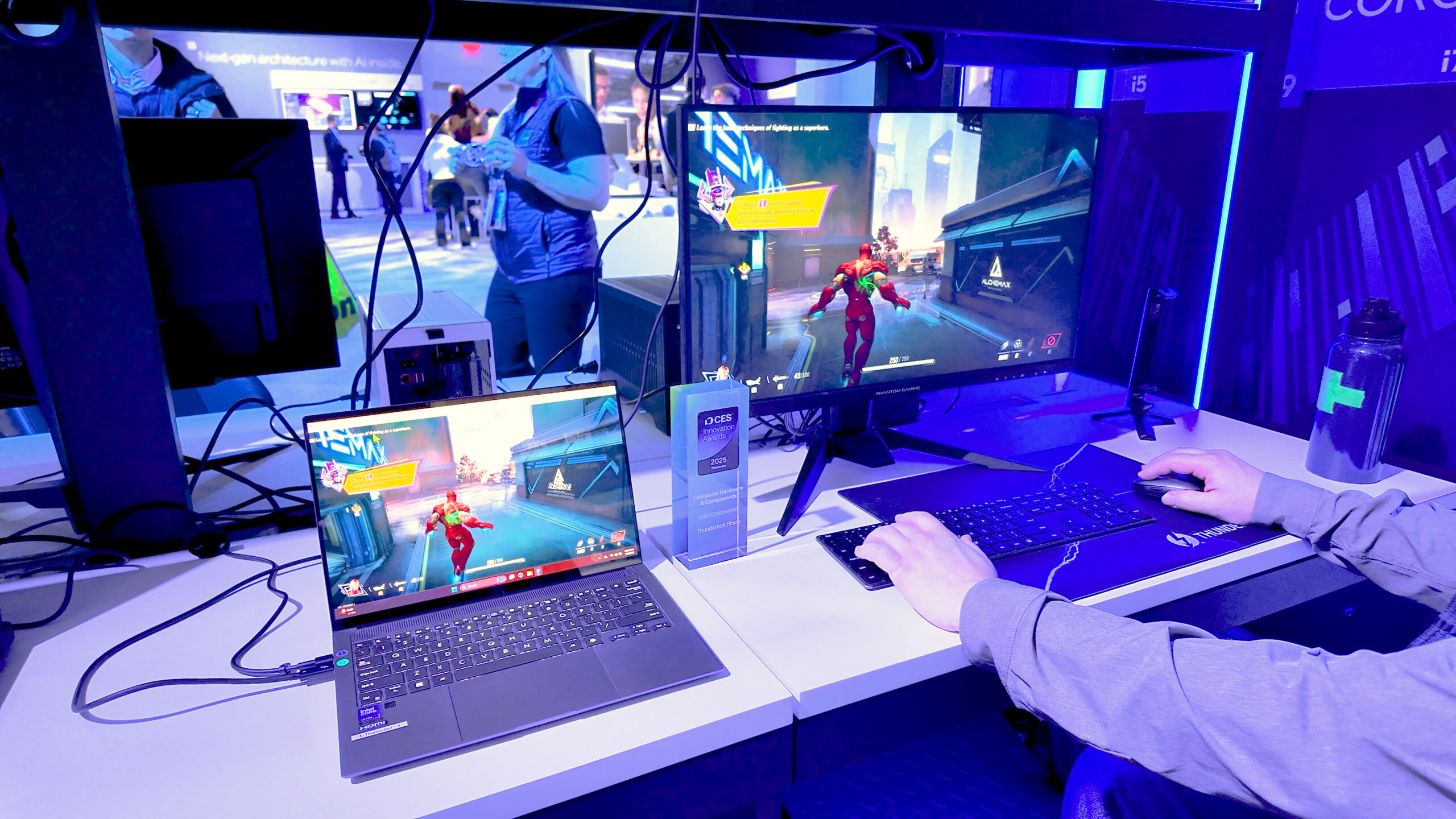 A person is playing a video game on a desktop computer and a laptop, both displaying the same game scene featuring a character running through a futuristic cityscape. The setup includes a keyboard, mouse, and a water bottle on the desk.
