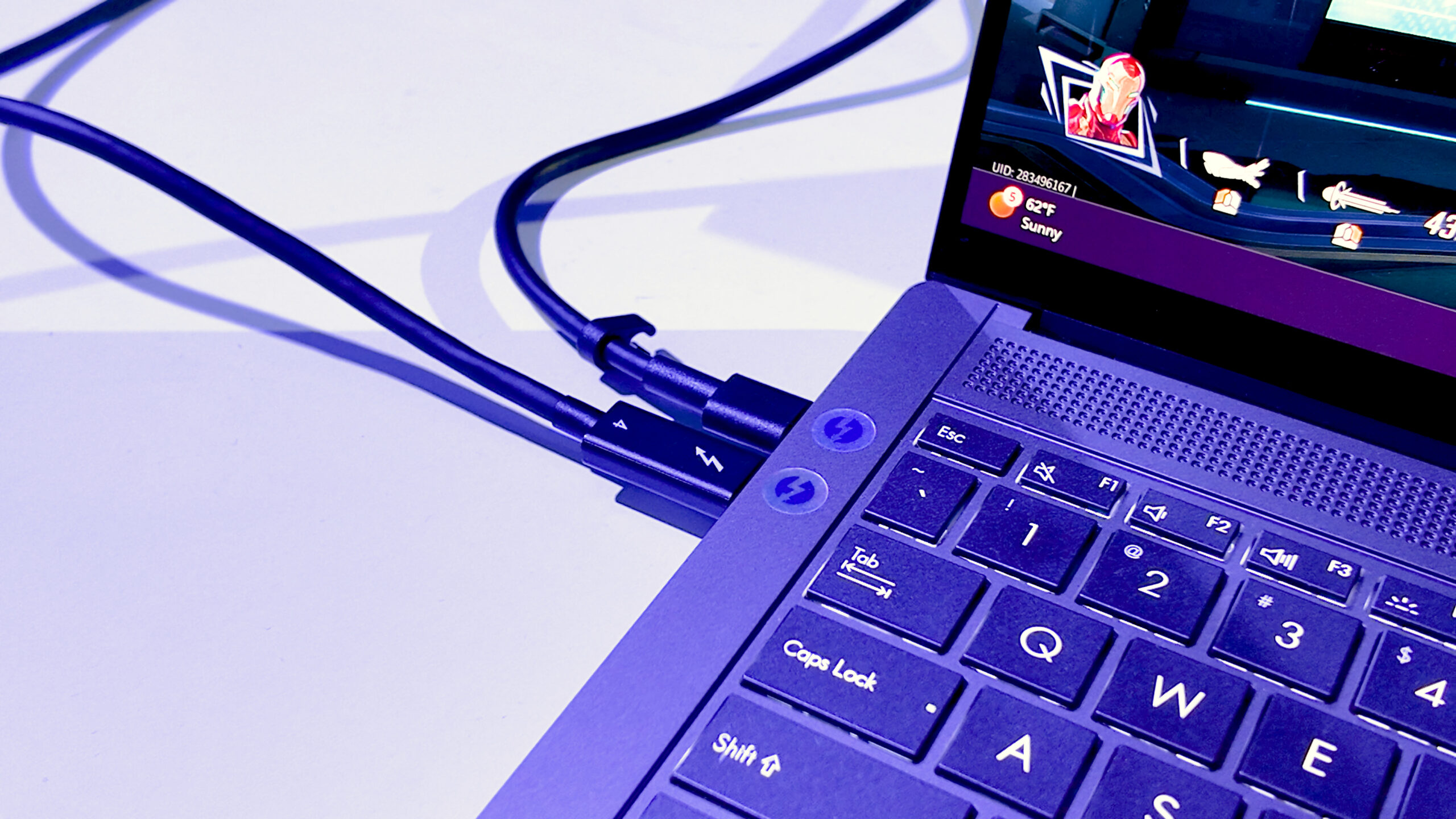 A close-up of a laptops keyboard and screen corner, with a focus on the USB-C and HDMI cables plugged in. The screen displays a video games interface with health and settings indicators. The lighting is blue-tinted.