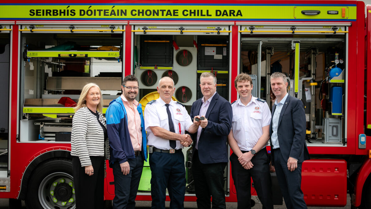 A group of six people, including a woman and five men, stand in front of a red fire truck with open compartments. The truck features Irish text. Two of the men shake hands while holding a ceremonial key.