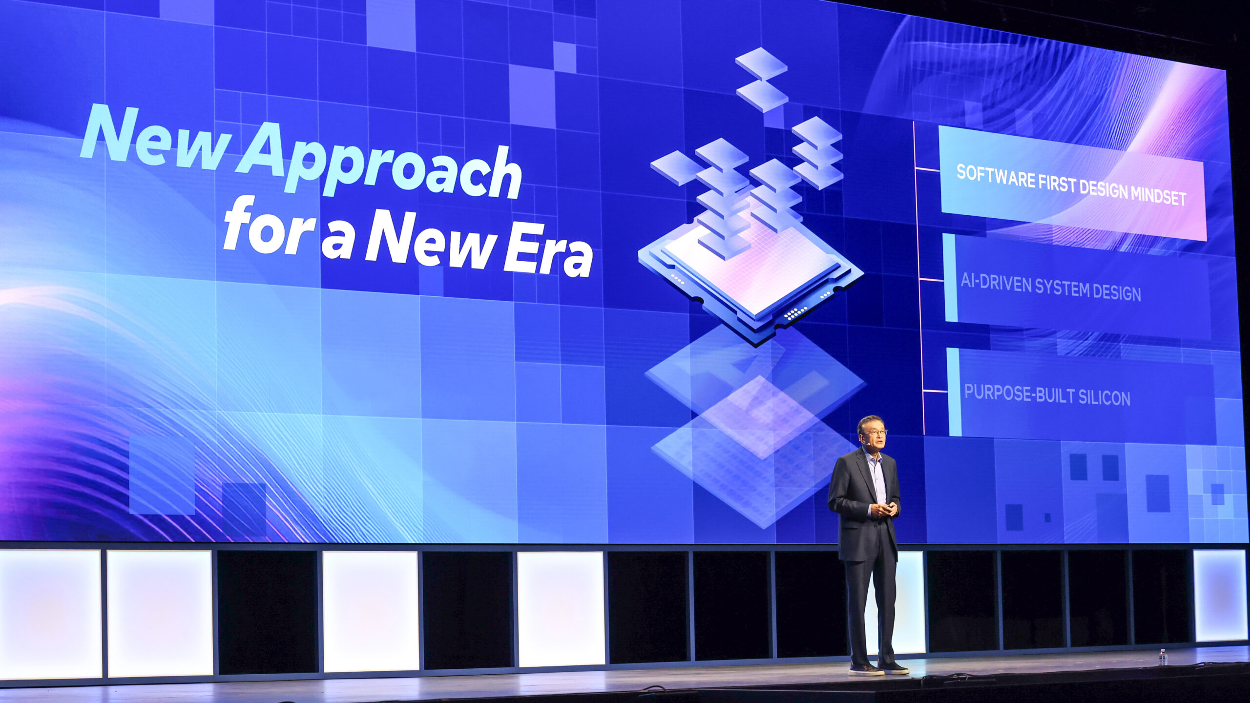 A person stands on a stage with a large blue screen behind them displaying text about a new design approach. The screen highlights Software First Design Mindset, AI Driven System Design, and Purpose-Built Silicon.