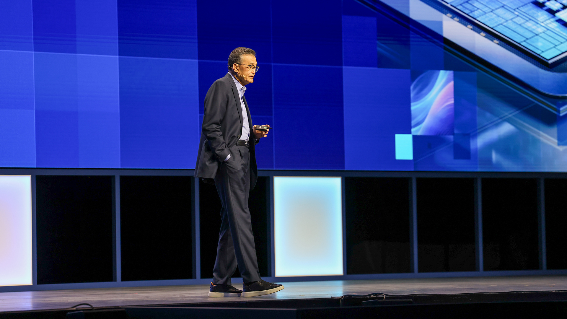 A person in a suit stands on a stage in front of a large blue digital screen, holding a small object.