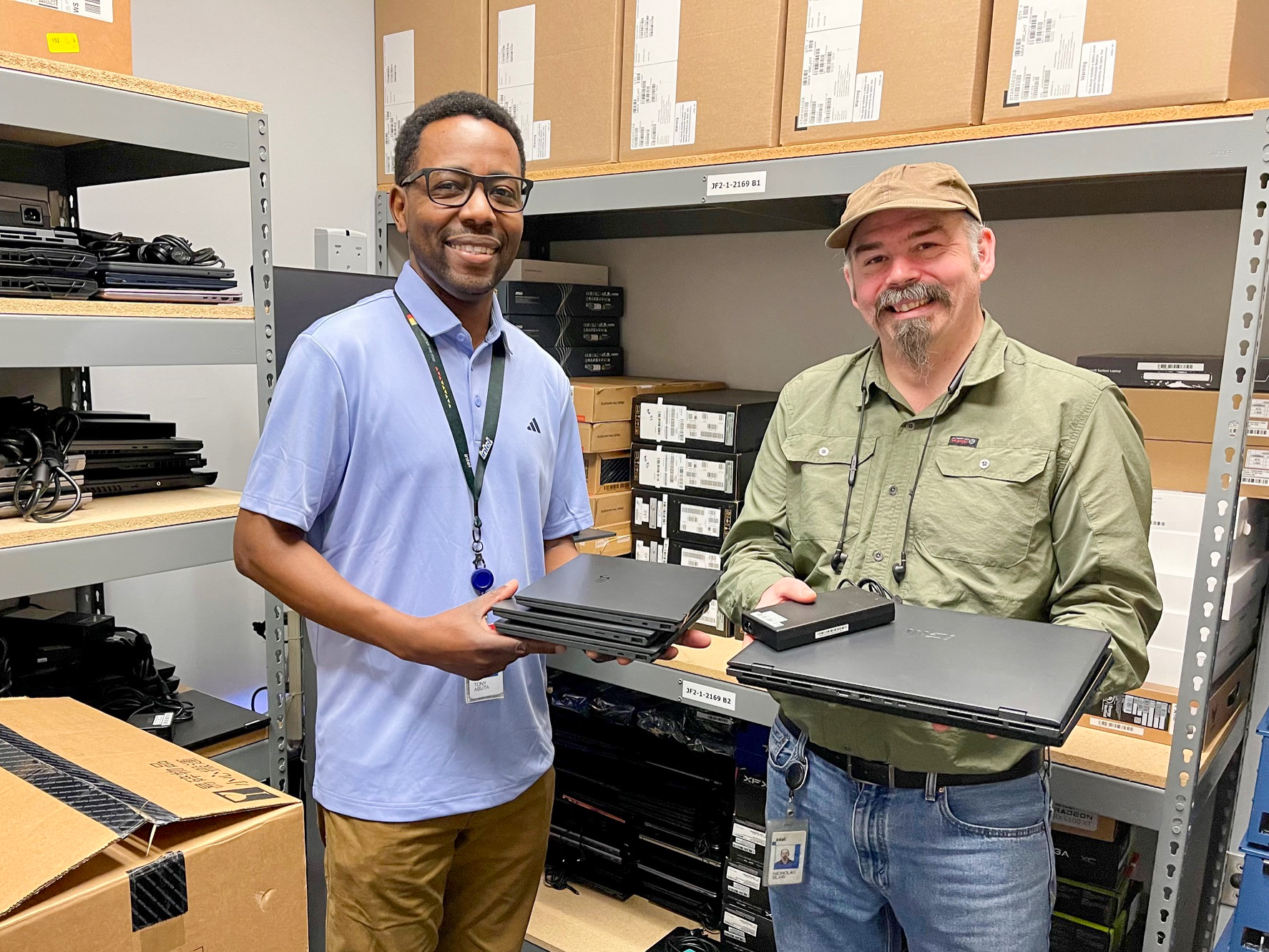 Dos hombres están de pie en un almacén lleno de cajas de cartón y dispositivos electrónicos. Ambos sonríen y sostienen pequeñas computadoras portátiles. Uno lleva una camisa azul claro y el otro, una camisa verde y una gorra beige.