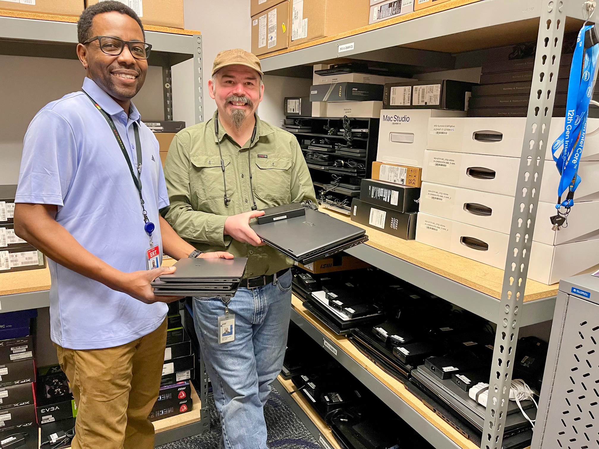 Dos hombres están de pie en un almacén con portátiles. A su alrededor, los estantes están llenos de diversos equipos tecnológicos y cajas. Uno lleva una camisa azul claro y el otro una camisa verde oliva con una gorra marrón. Ambos sonríen.