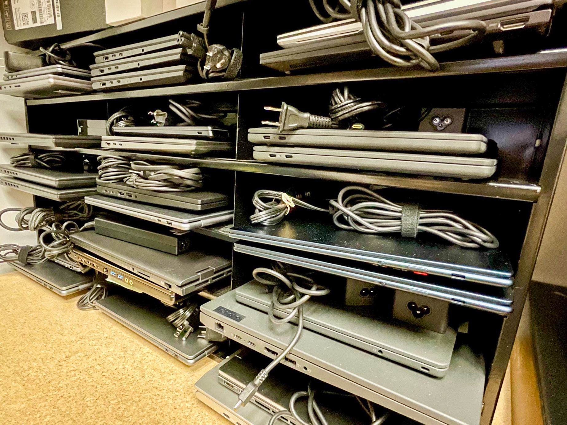 Una estantería llena de portátiles apilados y cables de alimentación organizados. Los estantes están abarrotados, y cada portátil tiene su cable perfectamente enrollado. La escena está muy iluminada, lo que realza el orden.