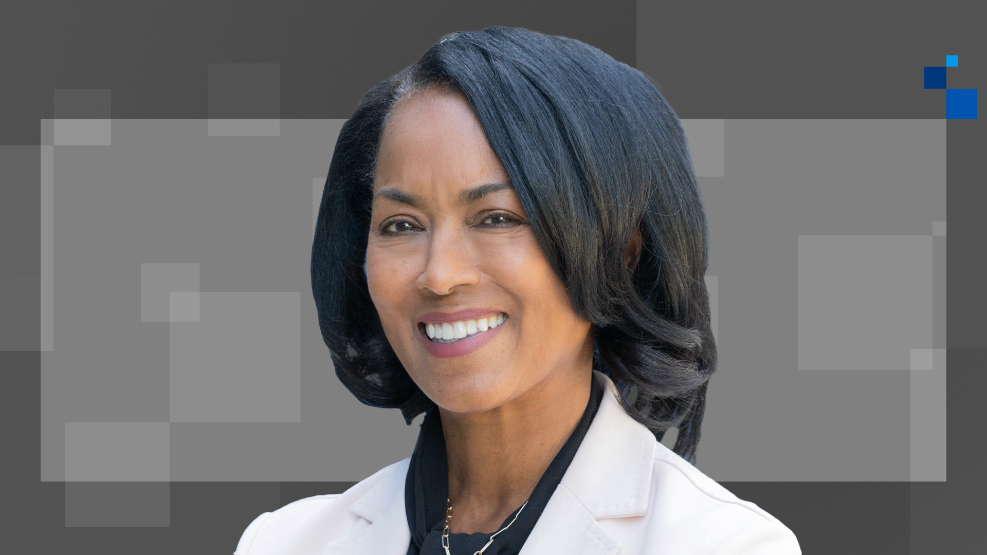 A woman with medium-length black hair smiles while wearing a light-colored blazer over a dark top. The background features overlapping gray and dark gray rectangles.