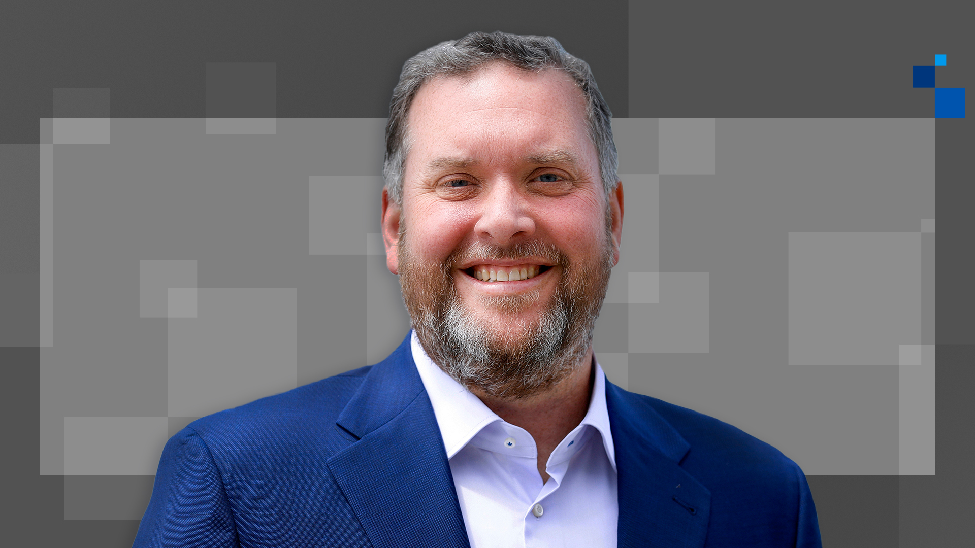 Smiling man with a beard wearing a blue suit and white shirt, standing against a gray background with abstract square patterns.