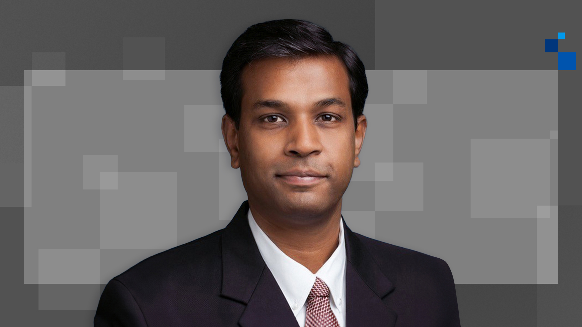 A person wearing a black suit, white shirt, and patterned tie is shown against a background with geometric gray shapes. The individual is facing forward, giving a formal and professional appearance.