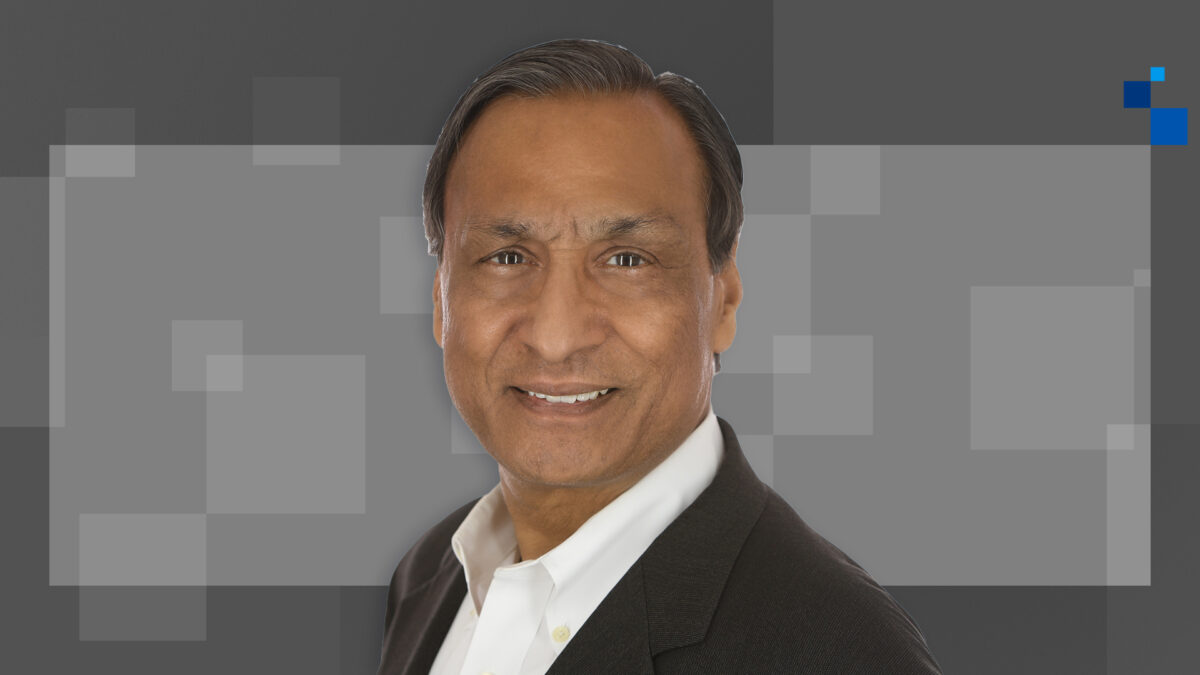 Smiling man in a black suit and white shirt against a gray geometric background with light squares and a blue corner detail.