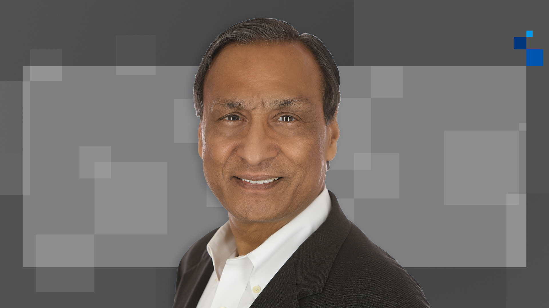 Smiling man in a black suit and white shirt against a gray geometric background with light squares and a blue corner detail.