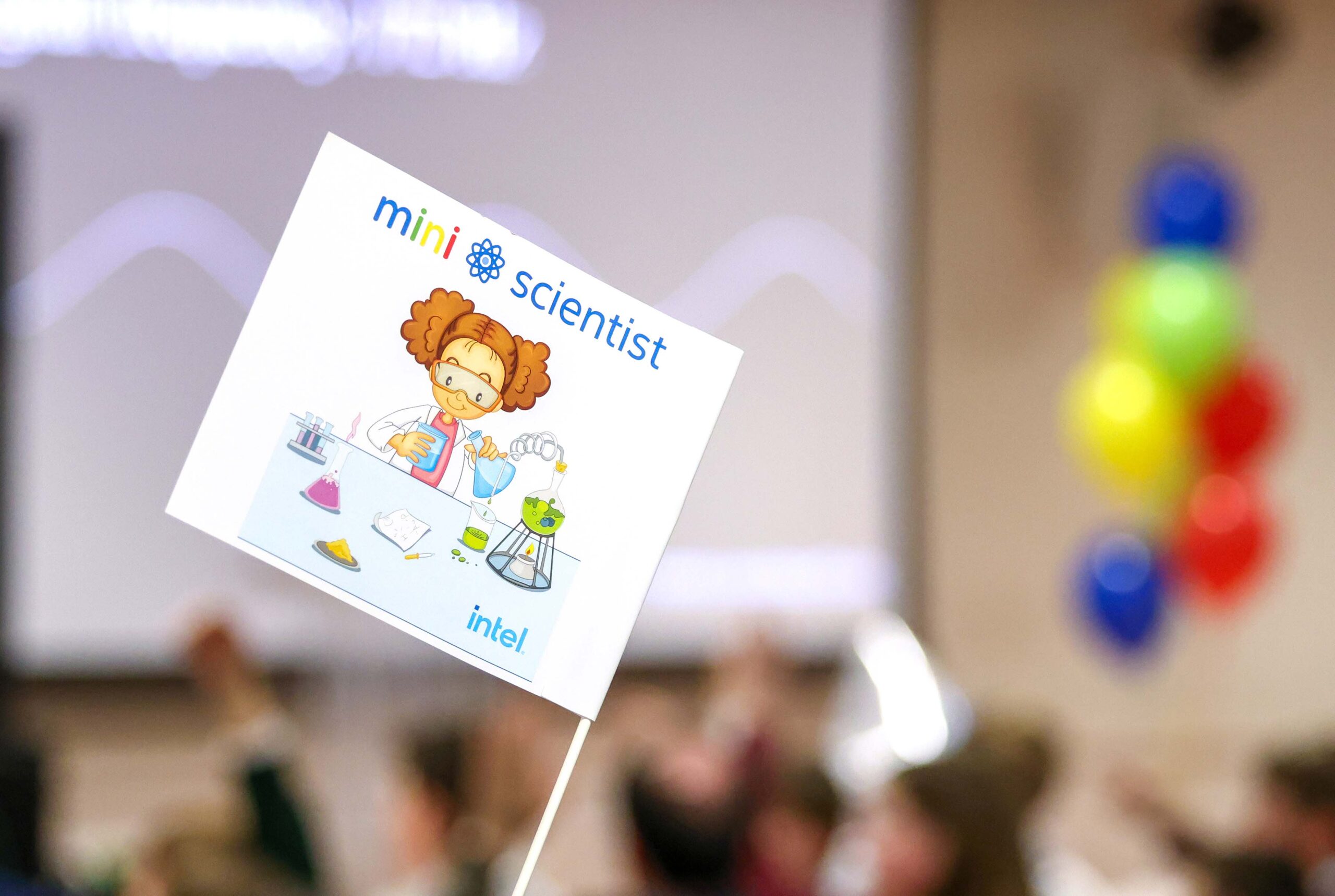 A small flag with a cartoon girl doing a science experiment, labeled mini scientist with an Intel logo. The background shows a blurred room with colorful balloons and an audience, suggesting a science event.