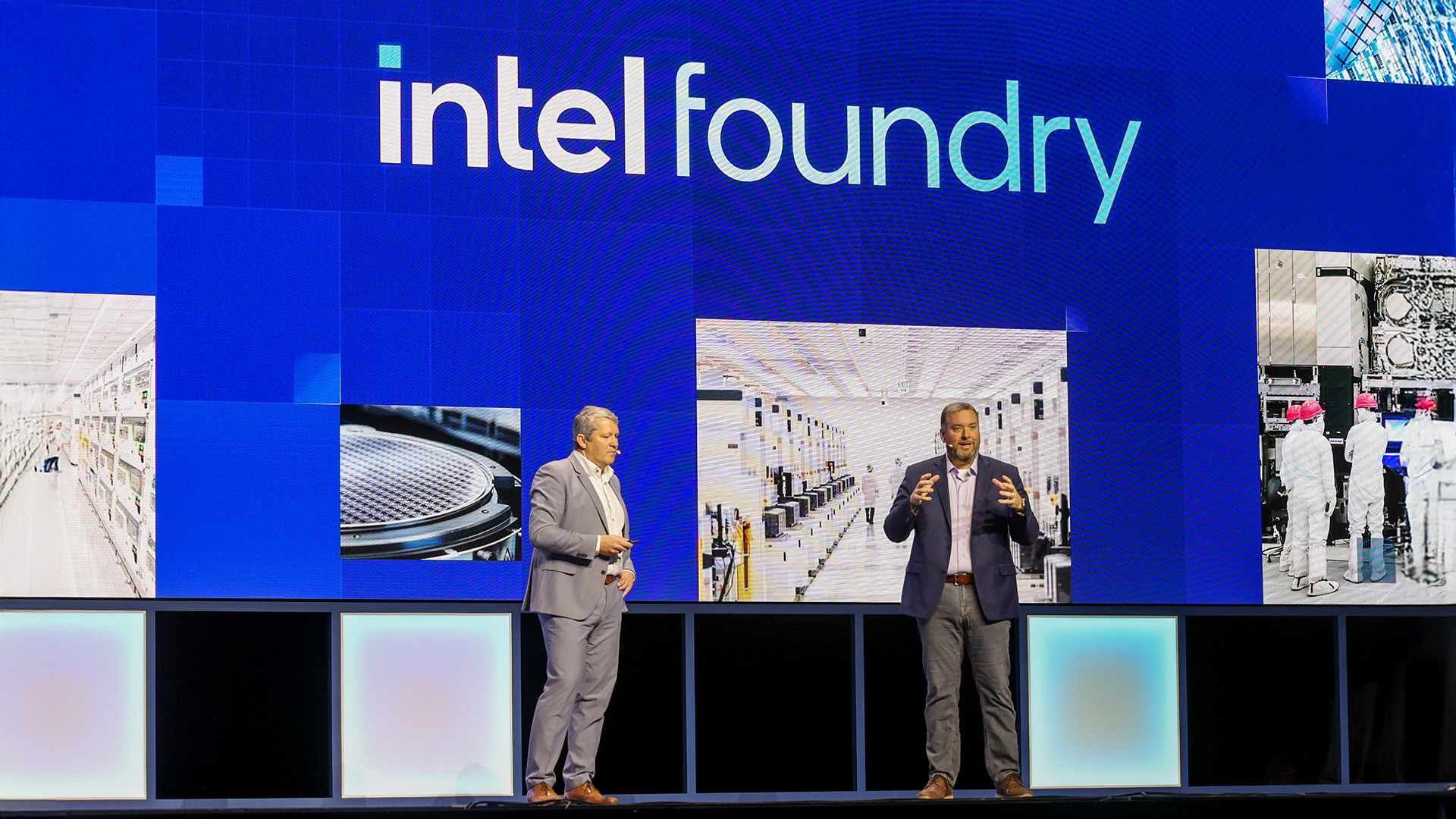 Two people stand on a stage under a large screen displaying intel foundry. The screen shows images of industrial scenes. Both are wearing suits, and one is gesturing while speaking.