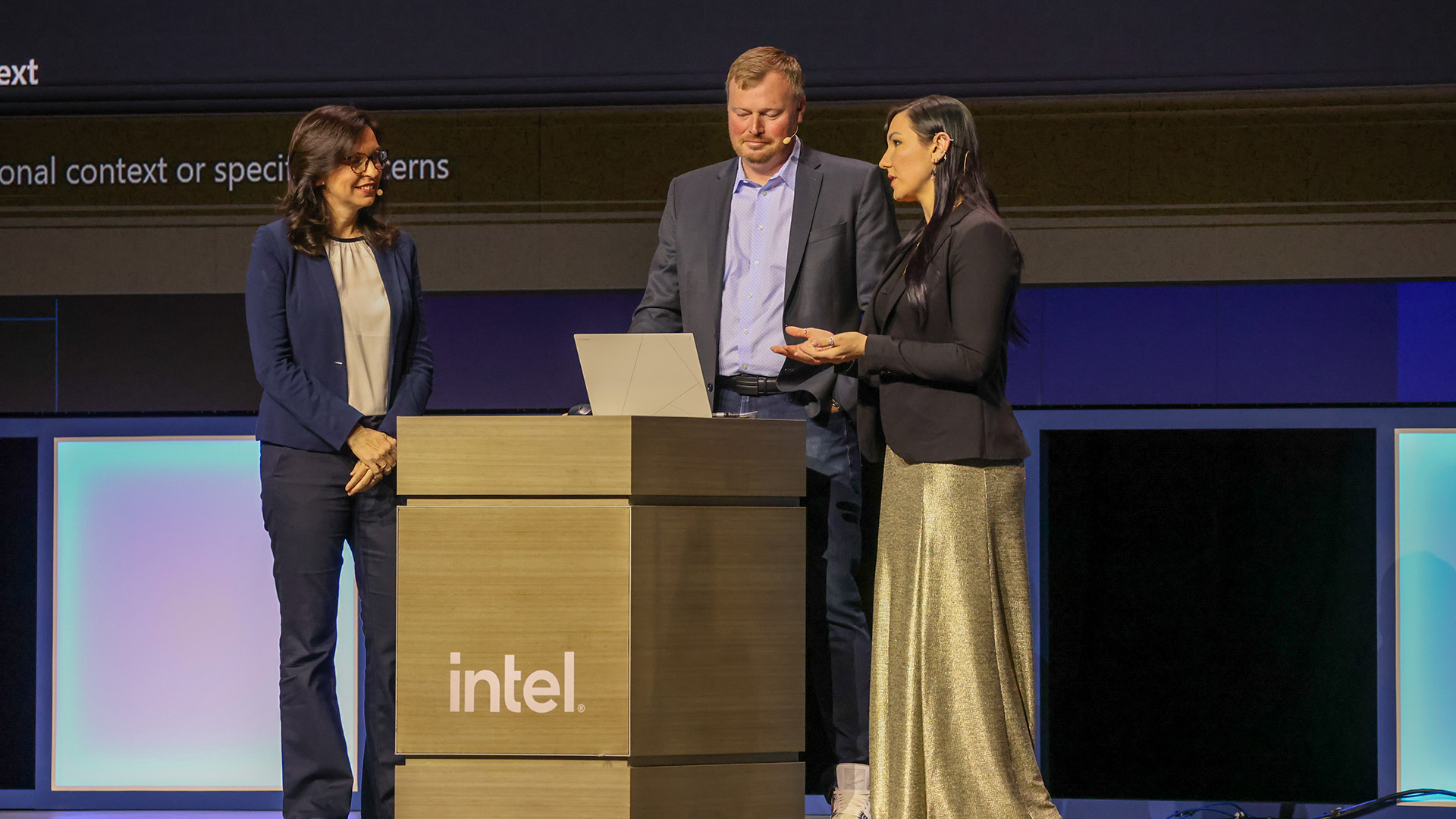 Three people stand on stage. Two women and a man stand behind a podium with an Intel logo. The woman on the right gestures while speaking. A laptop is open on the podium. The background is a large screen displaying partial text.