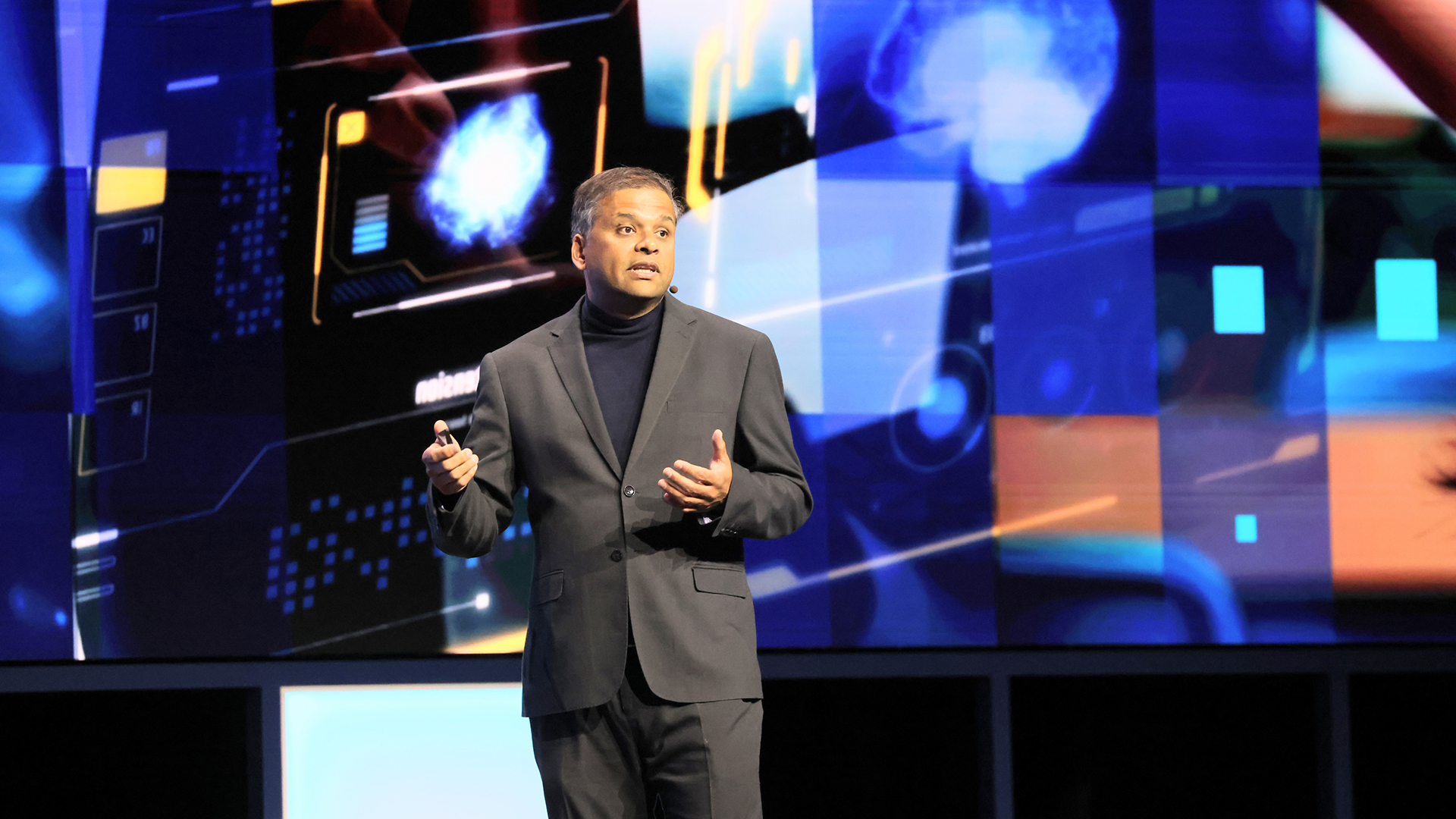 A man in a suit holds a microphone on stage, speaking in front of a large, brightly lit digital screen displaying abstract graphics and patterns.