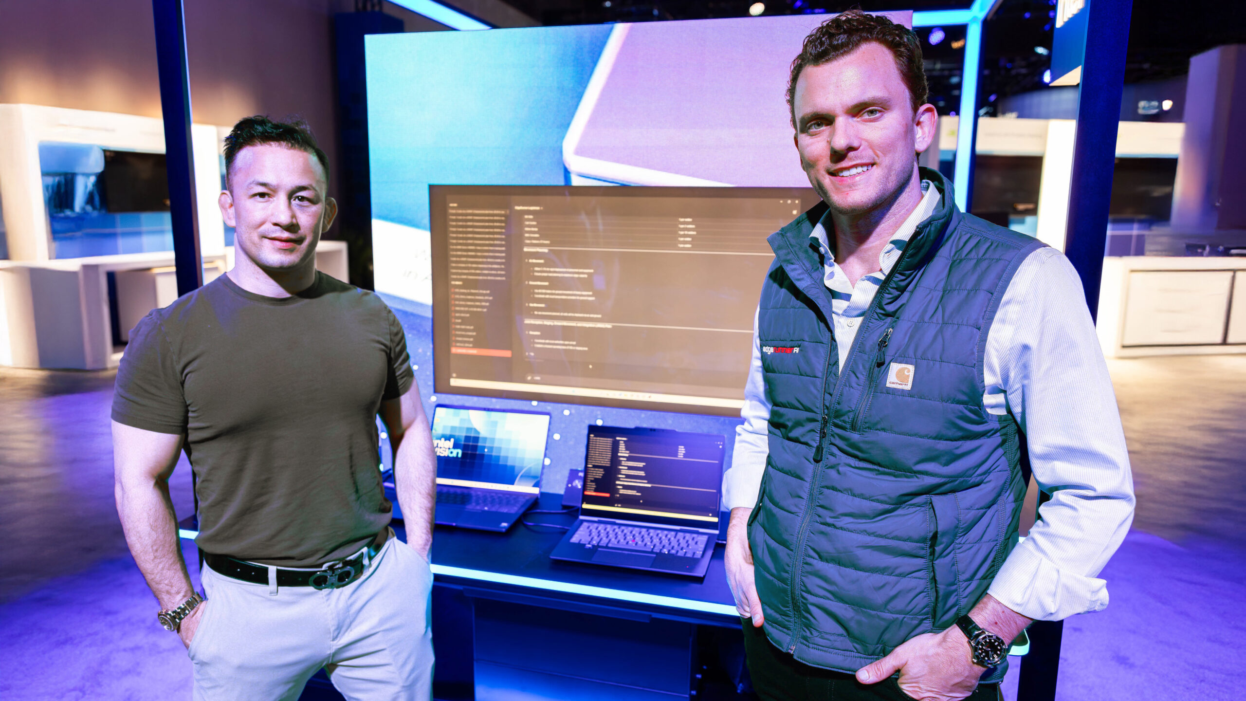 Two men stand in front of a tech display featuring a large monitor and two laptops. One wears a green t-shirt and jeans, the other a blue vest and shirt. The background is a tech exhibit with blue and purple lighting.