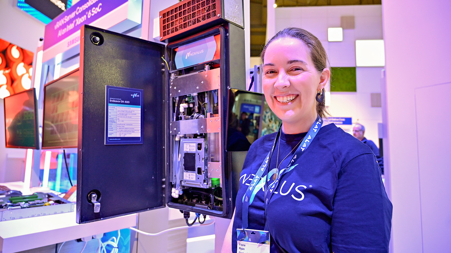 a smiling woman with her hair pulled back, wearing a blue t-shirt and a lanyard with badge, stands on a convention floor standing next to a piece of computer equipment